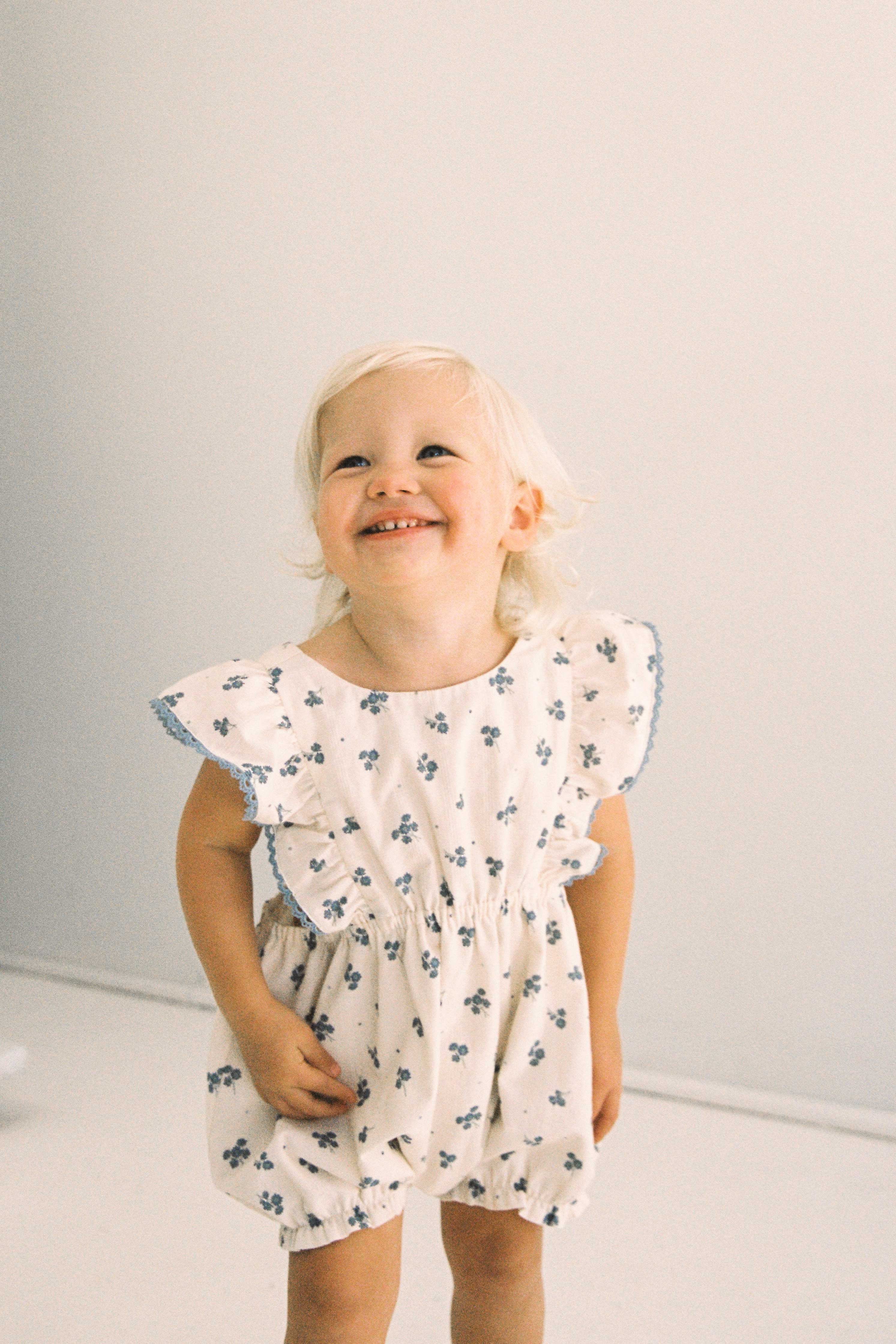 A young child with blonde hair smiles happily while looking up. The child is wearing the Daisy Romper Ditsy Floral from JUNI JUNIOR, a white dress made from textured cotton fabric, adorned with small blue flower patterns and ruffled sleeves. The ethically made outfit exudes charm against the plain, light-colored background.