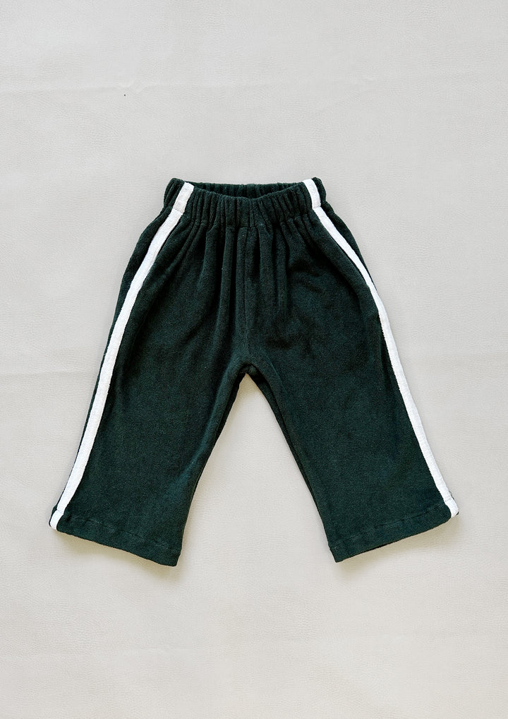 The Darcy Racer Pants Moss by TINY TROVE is a pair of toddler pants in a dark color with white side stripes and an elastic waistband, true to size. Crafted from premium terry towelling, the pants are laid out flat on a light gray background.