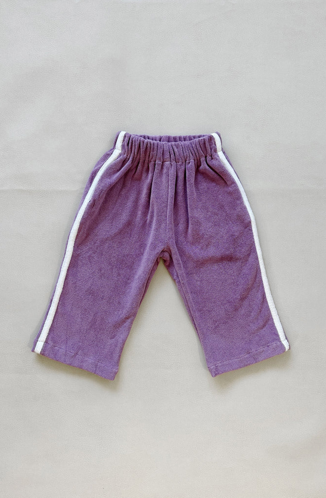 The Darcy Racer Pants Wisteria from TINY TROVE are crafted from soft, plush premium terry towelling. These purple pants feature an elastic waistband, white stripes running down the sides of each leg, and convenient pockets. They are displayed flat on a plain light-colored background.