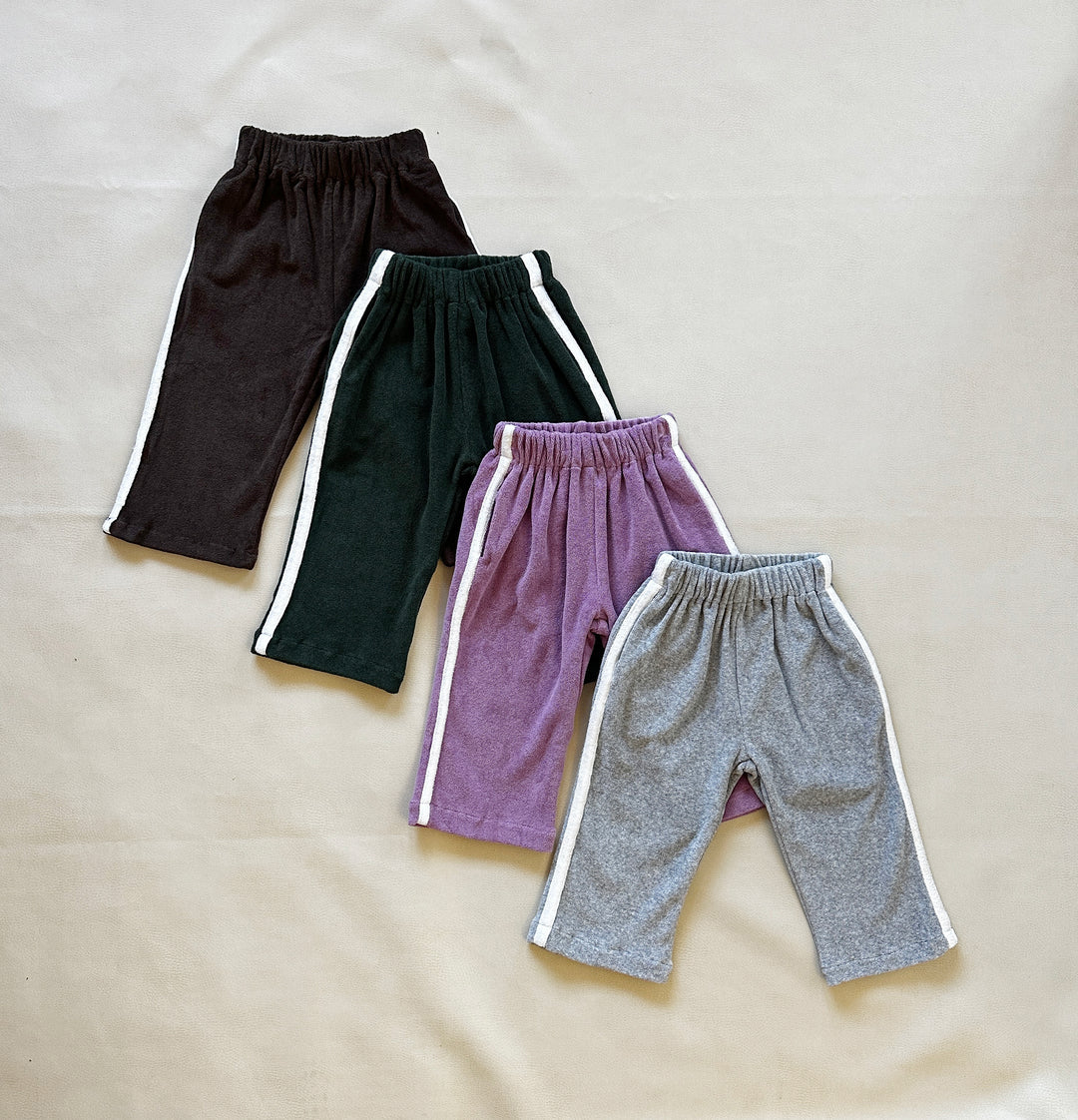 Four pairs of children's pants are laid out on a light-colored surface. The pants come in black, dark green, lavender, and gray, each with a stylish white stripe down the sides. Featuring elastic waistbands and crafted from premium terry toweling fabric, these soft Darcy Racer Pants Dark Chocolate by TINY TROVE also include convenient pockets for added functionality.