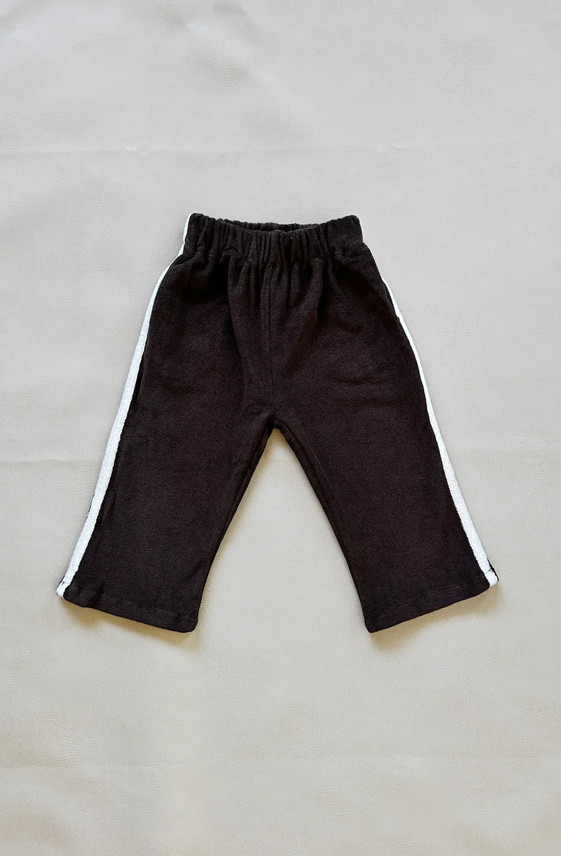 A pair of Darcy Racer Pants Dark Chocolate by TINY TROVE, featuring an elastic waistband, pockets, and white stripes running down the sides, made from premium terry towelling, laid flat on a light-colored surface.