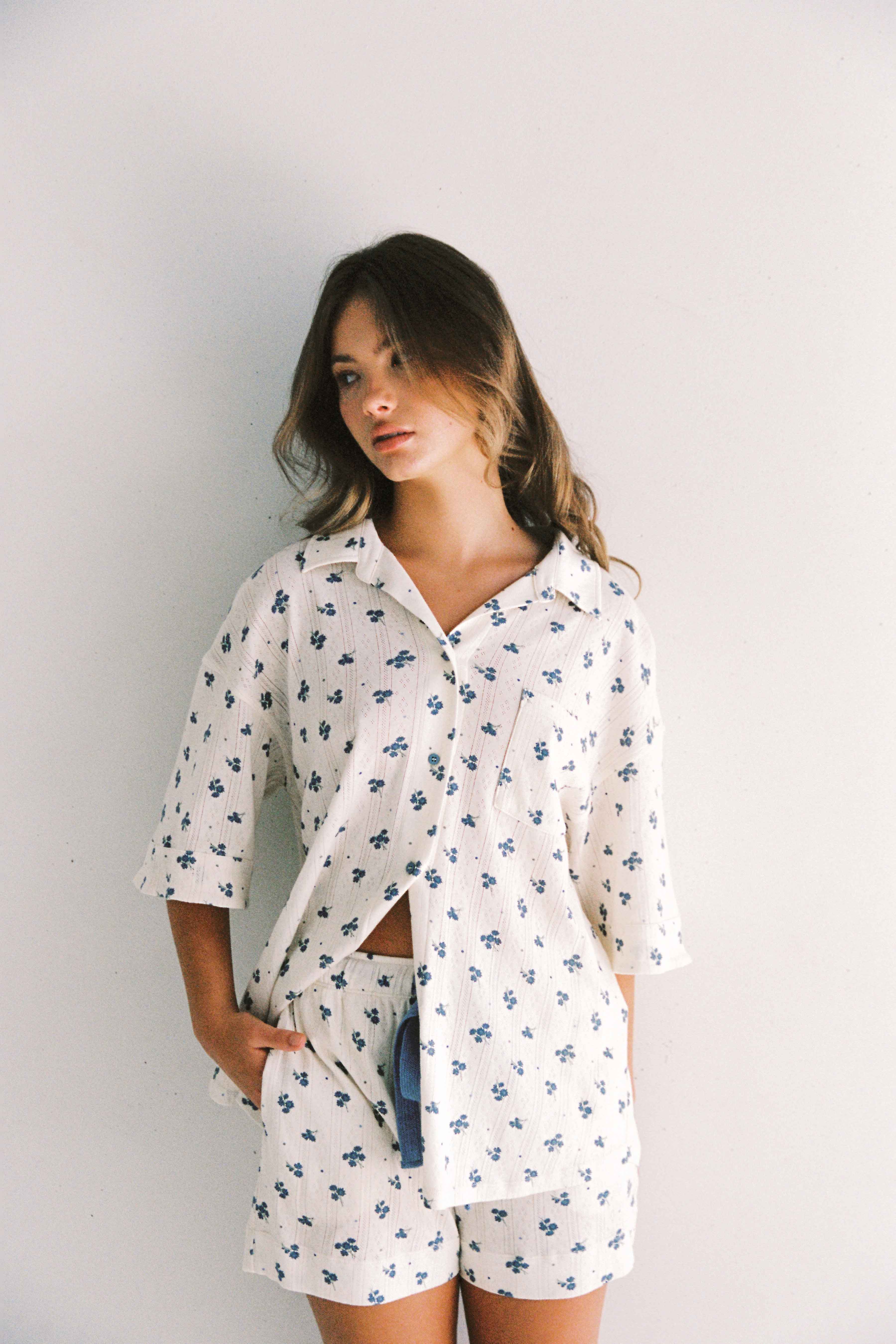 A young woman with long hair stands against a light gray wall. She is wearing the Daze Pyjama Set by JUNI, an ethically made outfit from China featuring a white button-up shirt and shorts with a blue floral pattern. Her left hand is in her pocket, and she gazes off to the side with a relaxed expression.