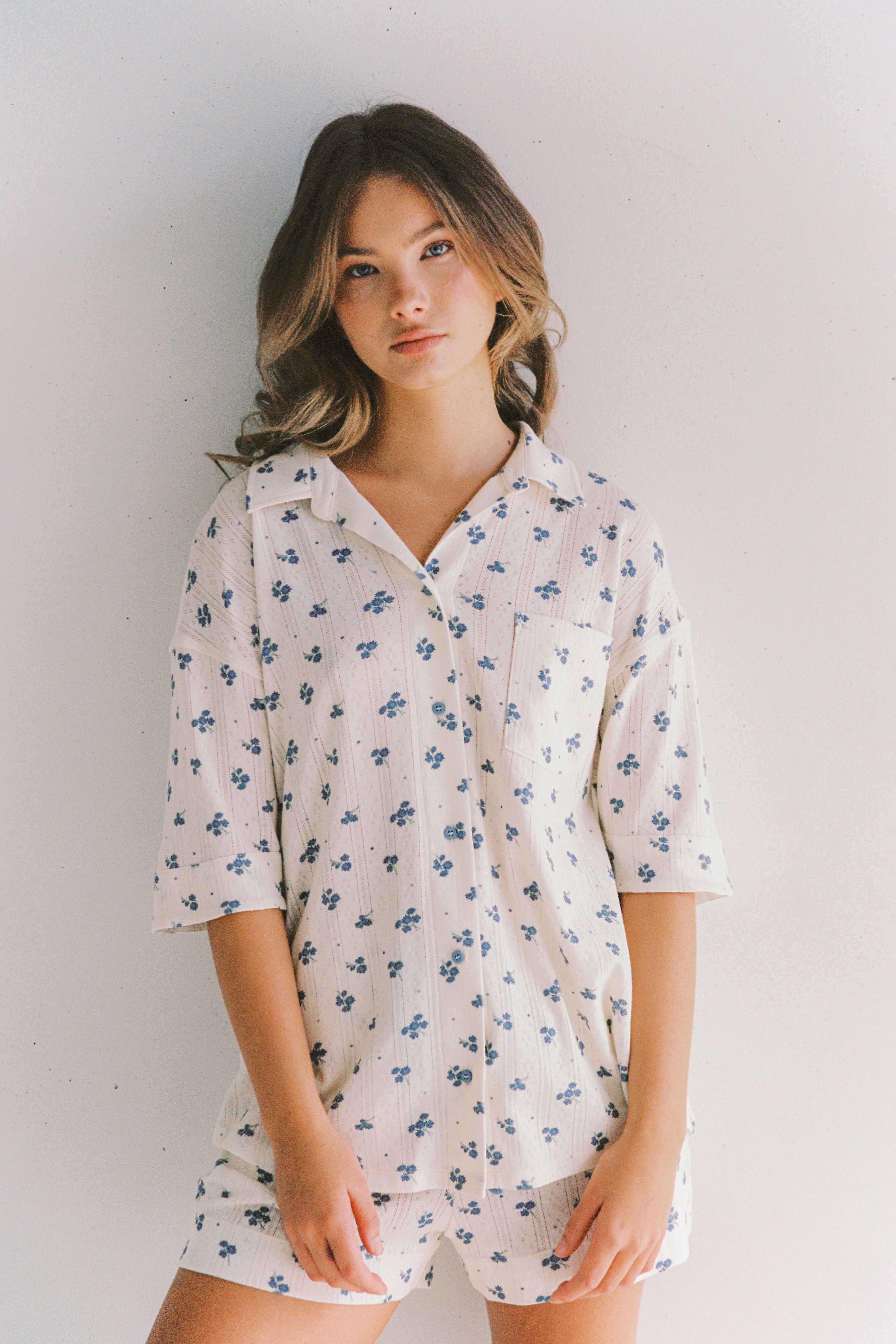 A person with long hair is standing against a white background, wearing the JUNI Daze Pyjama Set, which boasts a relaxed fit and is made from stretchy cotton featuring a blue floral print. With a relaxed expression and their hands casually placed at their sides, they perfectly showcase the comfort of this ethically made in China sleepwear.