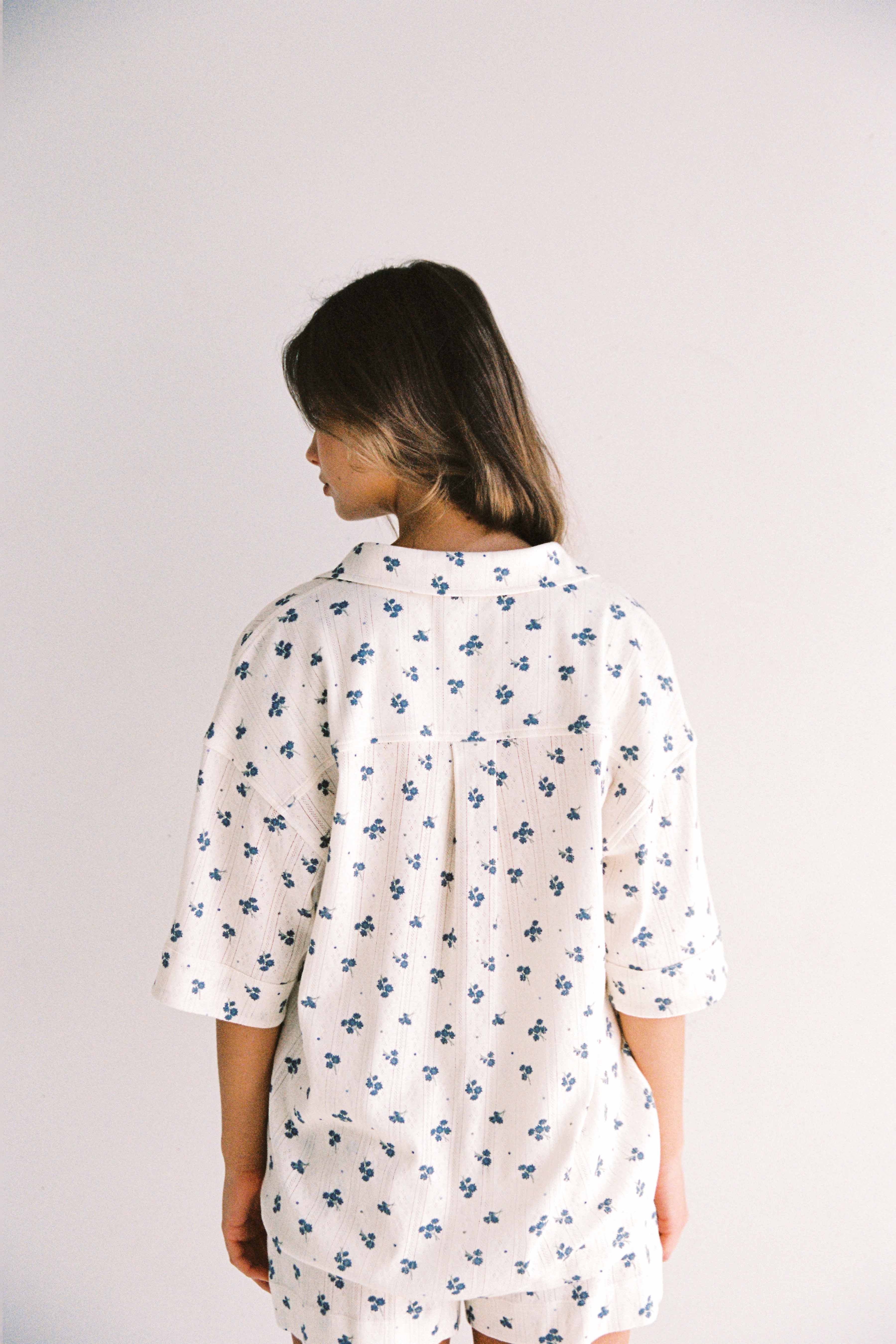 A person stands facing away from the camera against a plain white background, dressed in a Daze Pyjama Set by JUNI. They are wearing a white shirt adorned with blue floral prints and sleeves that reach just below the elbows. Their dark hair falls loosely around their shoulders, complementing the relaxed fit pyjama set made from stretchy cotton.