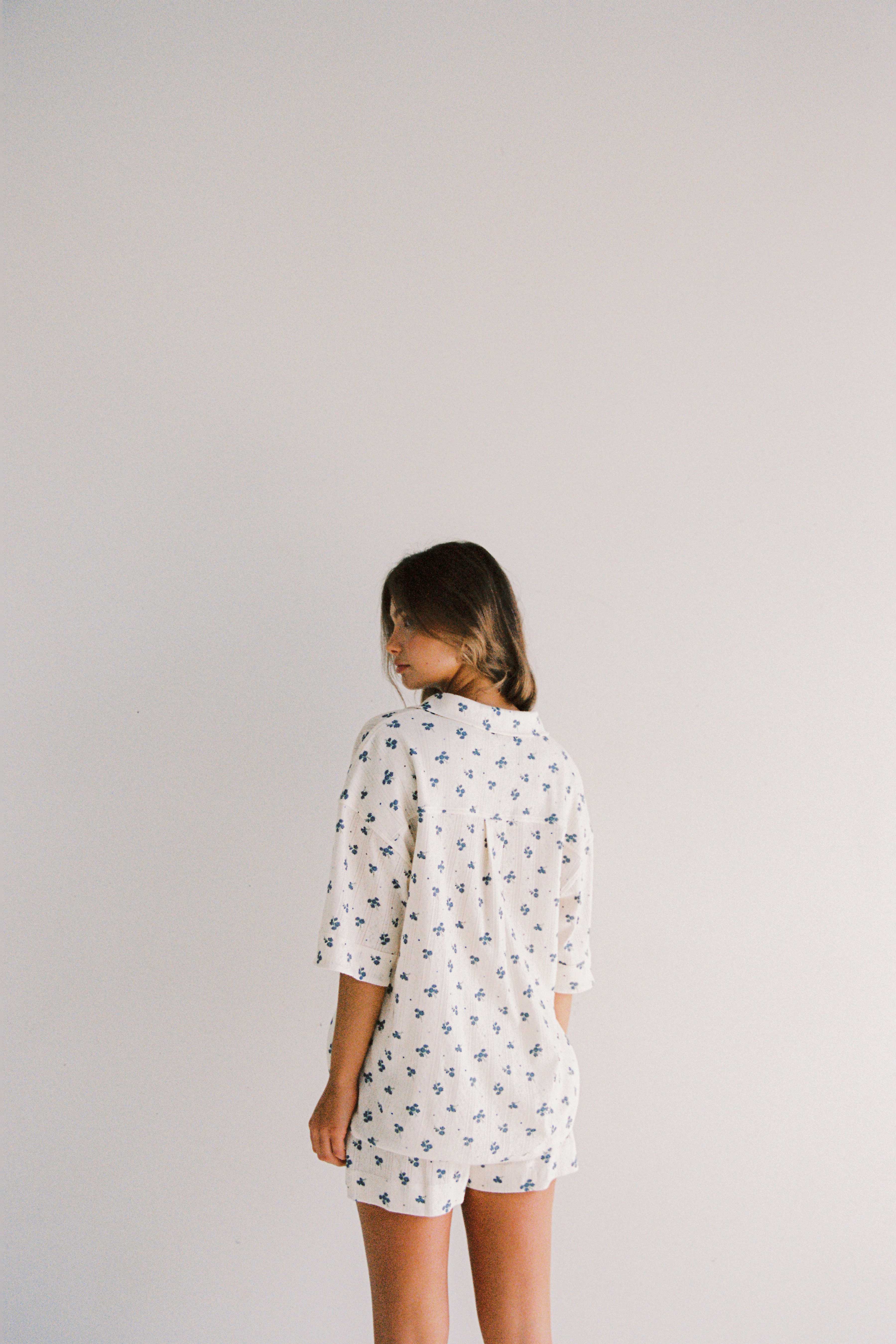 A person with long hair stands against a plain white background, wearing the Daze Pyjama Set by JUNI. Ethically made in China, this loose-fitting, light-colored outfit features a delicate small blue floral pattern. The person is slightly turned away, looking over their shoulder while donning the relaxed fit pyjama set.