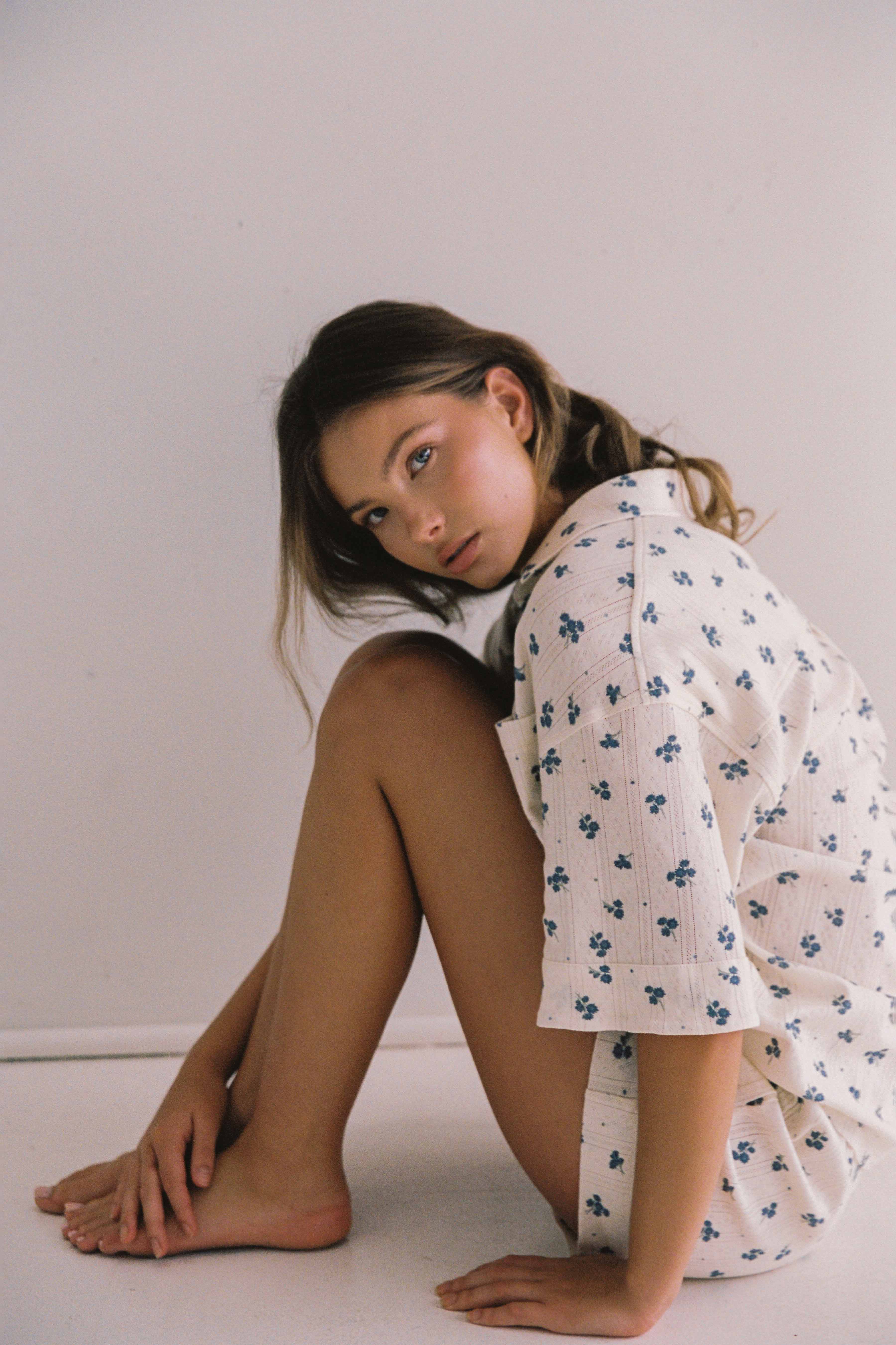 A person with long hair is seated on the floor against a plain white background, wearing the Daze Pyjama Set from JUNI. The cream-colored, relaxed fit pyjama, adorned with a blue floral pattern, is made from stretchy cotton and ethically crafted in China. With one arm resting on their knees, they look at the camera with a relaxed expression.