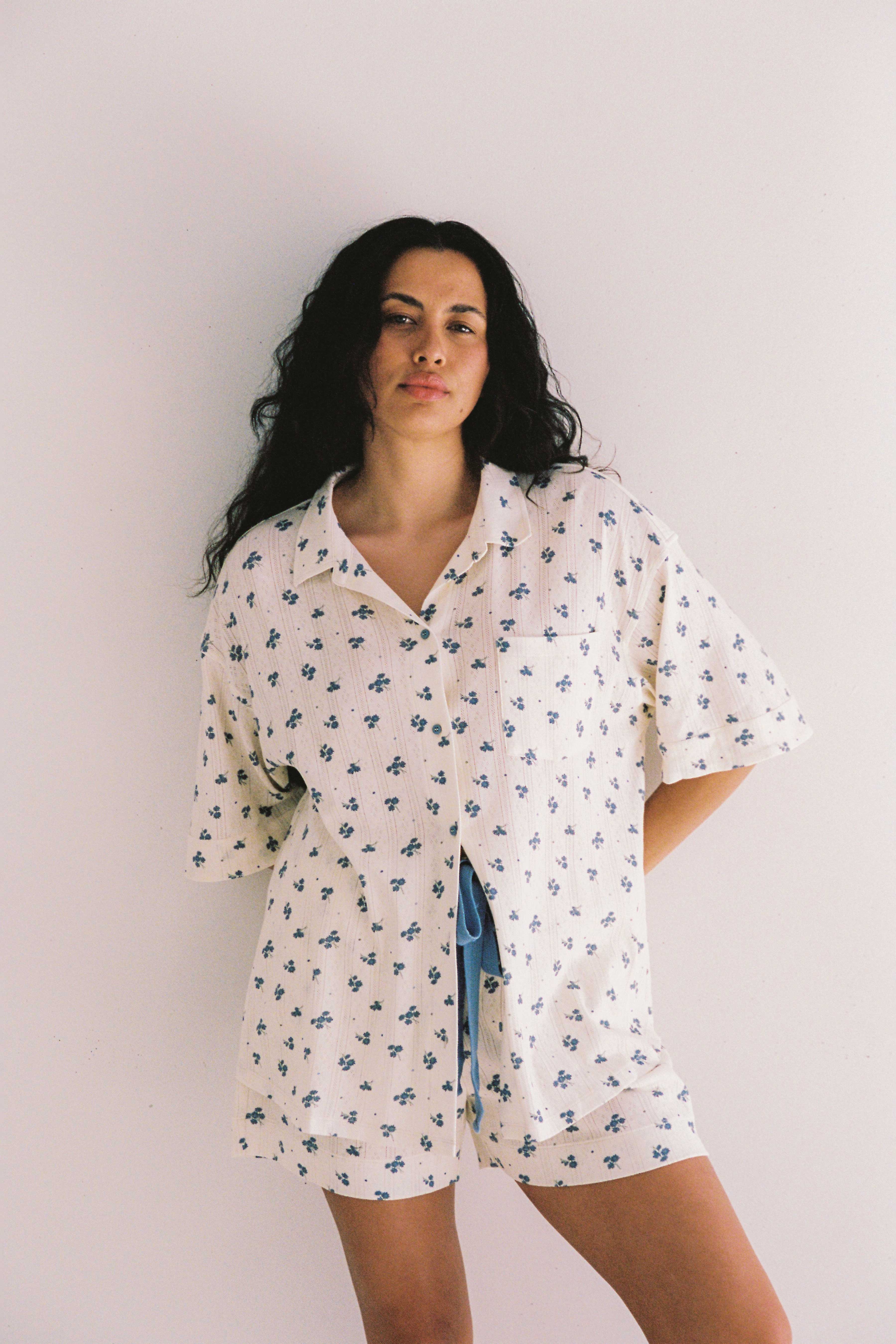 A person with long, wavy hair stands against a plain white background, dressed in the Daze Pyjama Set from JUNI. The loose-fitting, short-sleeve top features a blue floral pattern and is ethically made in China. They have a relaxed expression as their left hand rests partially in their pocket.