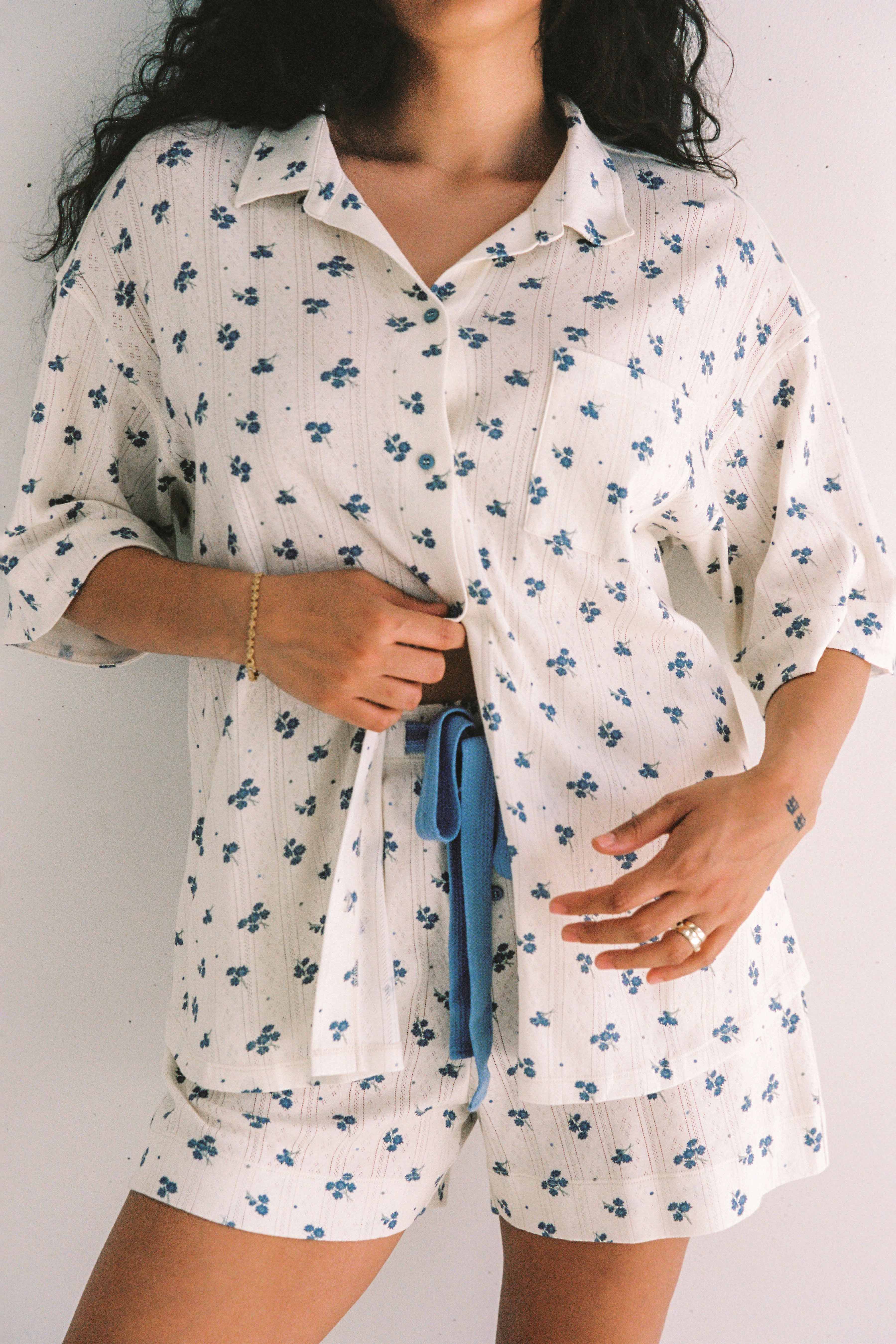 A person is wearing the Daze Pyjama Set by JUNI, featuring a white fabric adorned with small blue floral patterns and crafted from stretchy cotton. The top has short sleeves and buttons down the front, while the shorts are equipped with a coordinating blue drawstring. Ethically made in China, this relaxed fit pyjama offers exceptional comfort as the individual gently holds the top near their waist.