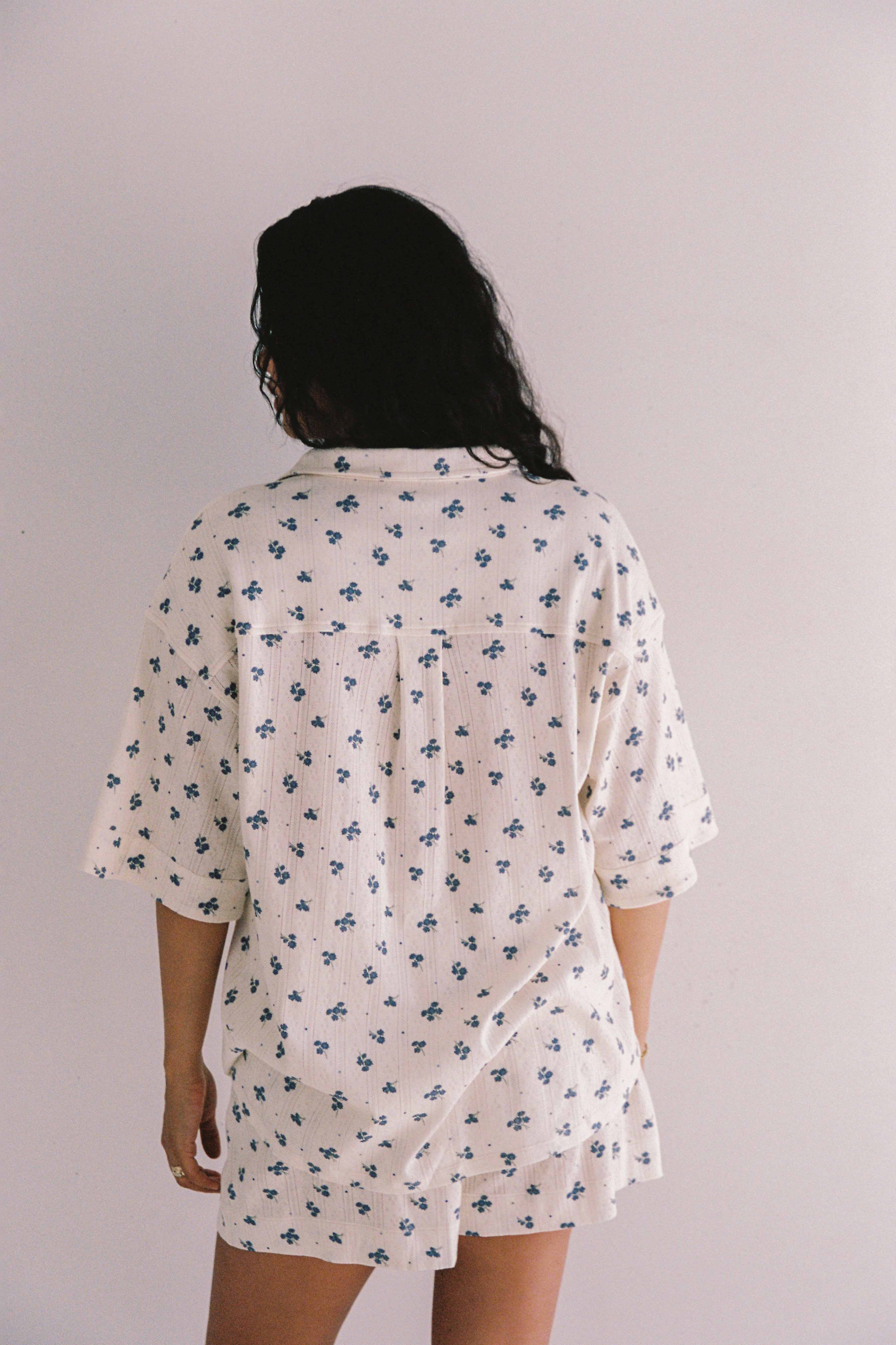 A person with long, dark hair stands with their back to the camera, wearing the Daze Pyjama Set from JUNI, featuring a relaxed fit and a blue floral pattern. This ethically-made in China outfit includes a white shirt and matching shorts against a plain, light-colored background.