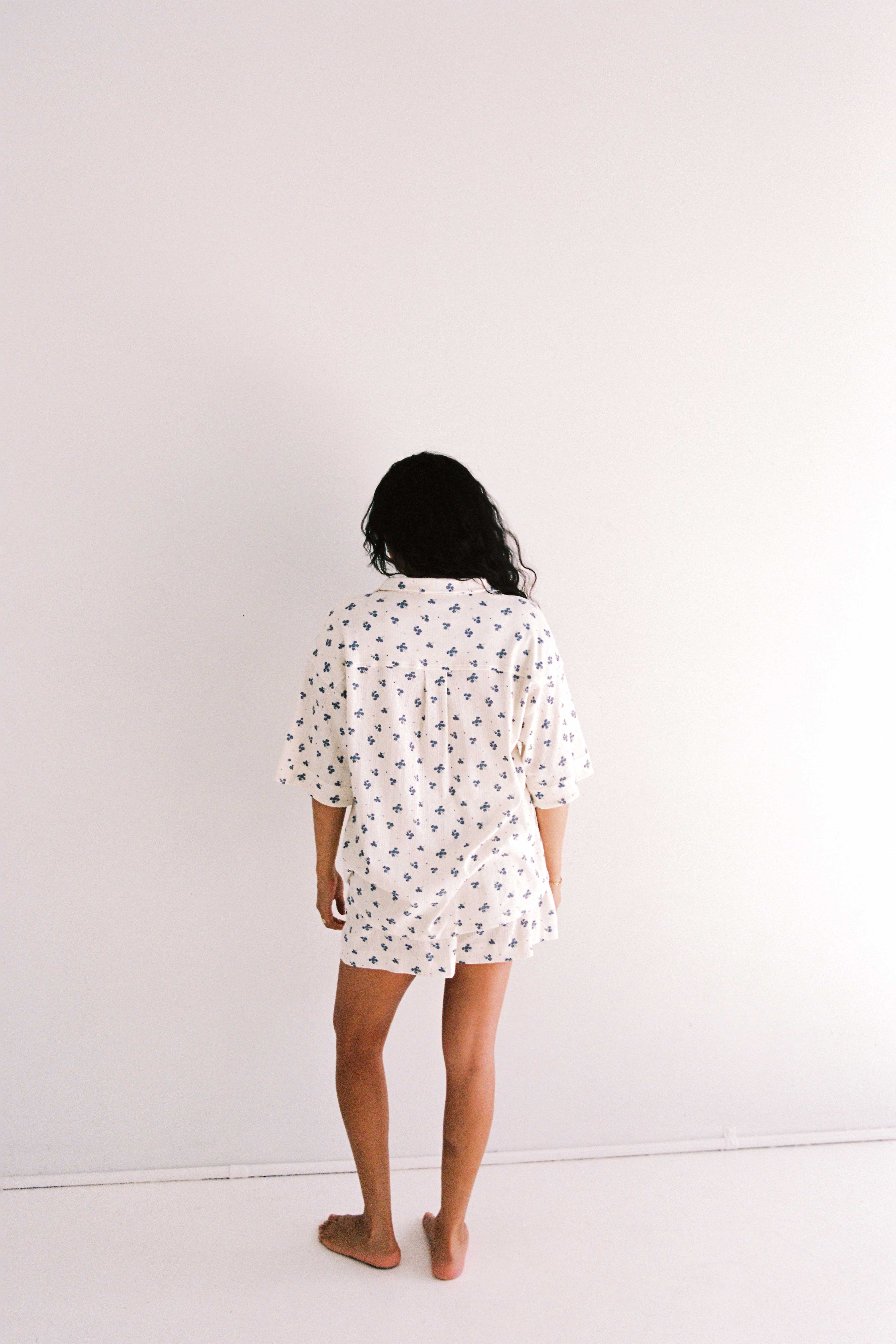 A person with long, dark hair stands barefoot facing a plain white wall. They are wearing the Daze Pyjama Set by JUNI, which is ethically made in China and features a relaxed fit with a small, dark pattern. Their head is slightly turned to the right, and one leg is slightly bent. The overall setting is minimalistic.