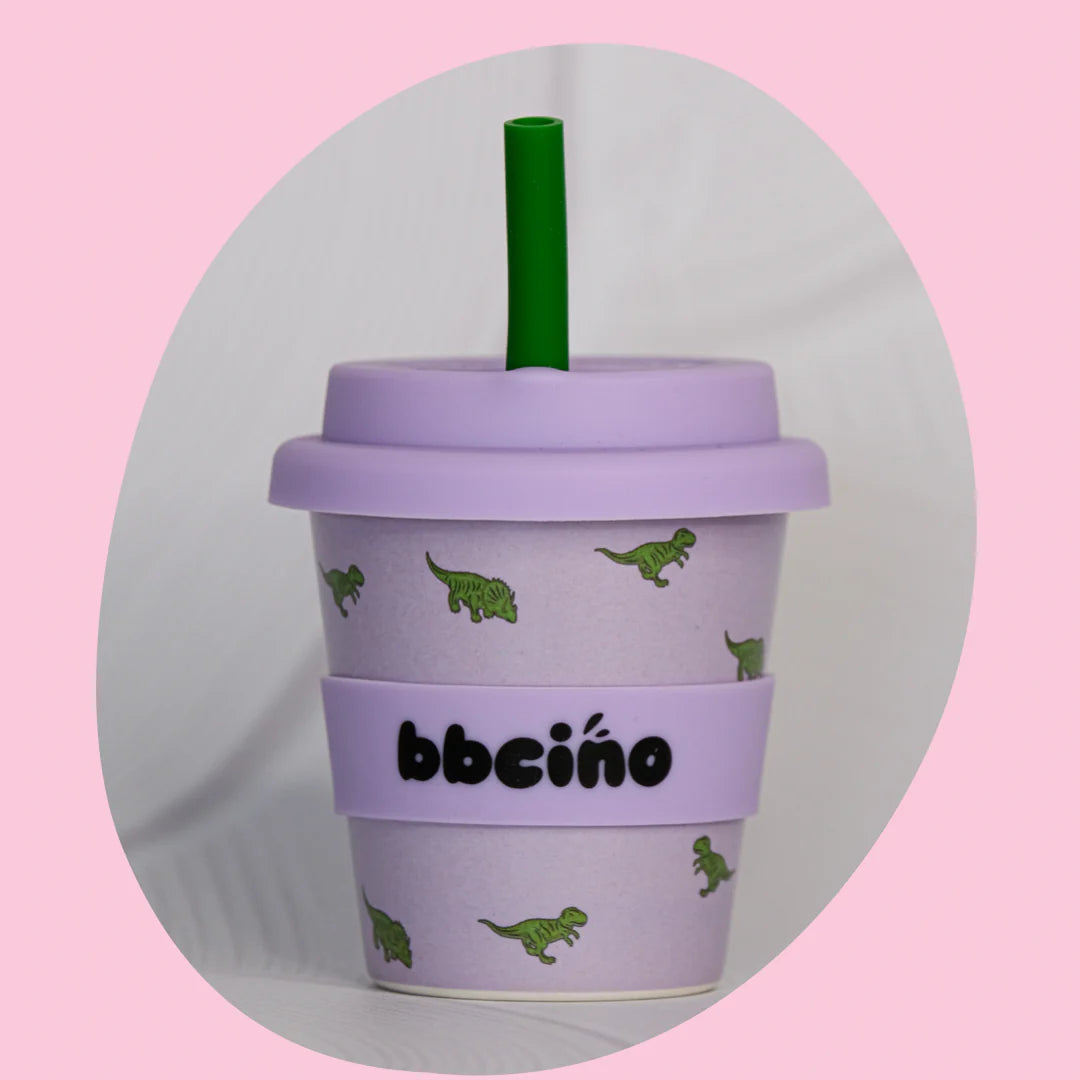 The Reusable Babycino Cup 120ml Dino-mite by BBCINO is a pastel-colored, sustainable babycino cup decorated with small green dinosaur illustrations. The light purple cup comes with a matching lid and a reusable silicone straw. Made from bamboo fibers, the sleeve features "BBCINO" in black. The image is set against a light pink background.