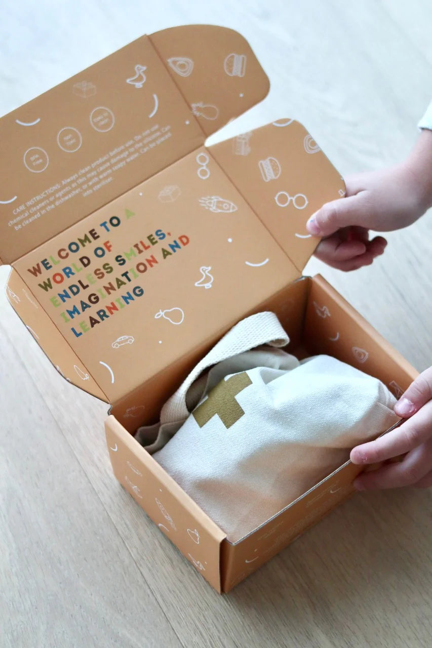 A pair of hands opening a cardboard box with playful illustrations. The inside flap reads, "Welcome to a world of endless smiles, child's imagination, and learning." Inside, there's a cloth bag adorned with a golden cross. This LITTLE DROP Doctor Set rests on a light wooden surface.