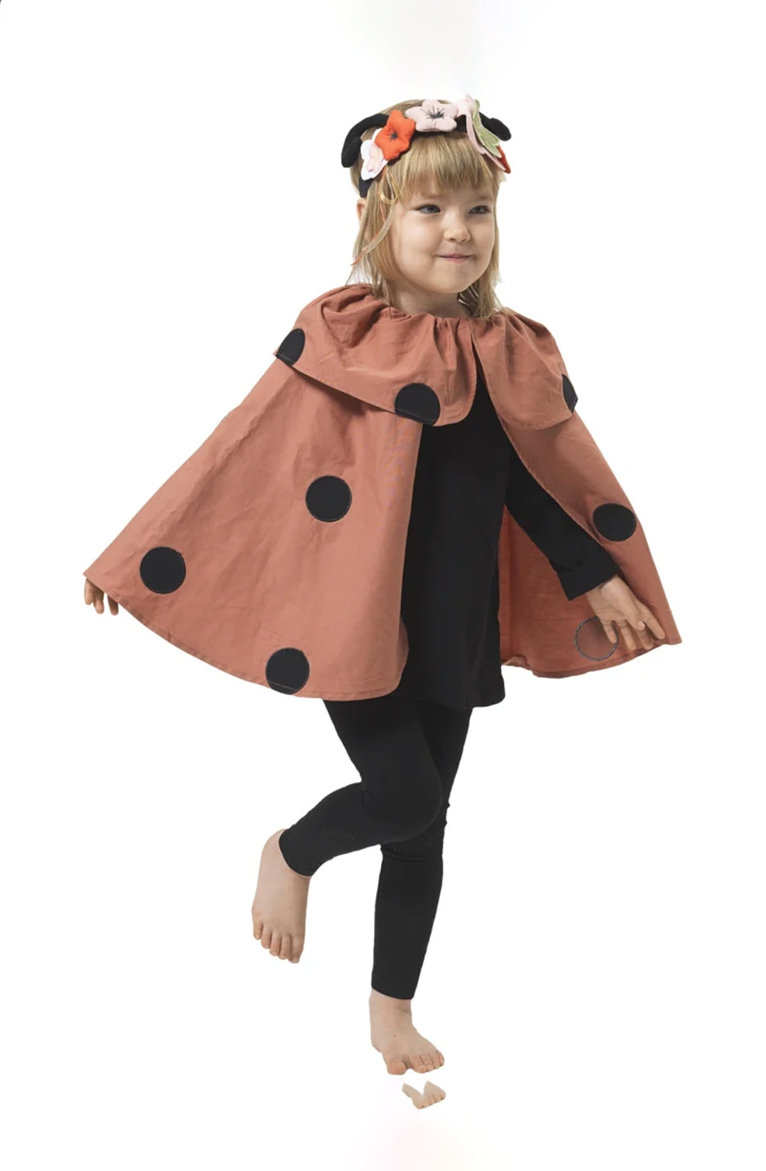 A young child, dressed in FABELAB's Dress Up Ladybug Set featuring a red cape with large black dots and a headband with antennae and flowers, wears an organic cotton black outfit underneath. Standing on one leg against a plain white background, the child smiles brightly, embracing children's imagination.