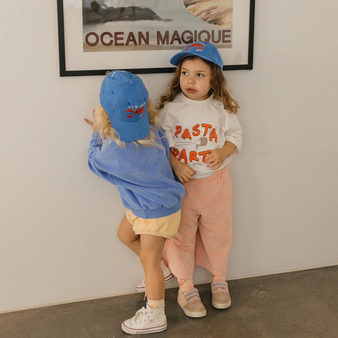 Two young children in vibrant outfits and blue Lobster Caps by GOLDEN CHILDREN, each featuring an adjustable brass clasp, stand before a framed picture on the wall. One child sports a blue sweater paired with shorts, while the other wears a sweatshirt adorned with "PASTA PARTY" text and peach pants. They seem to be talking or playing together.