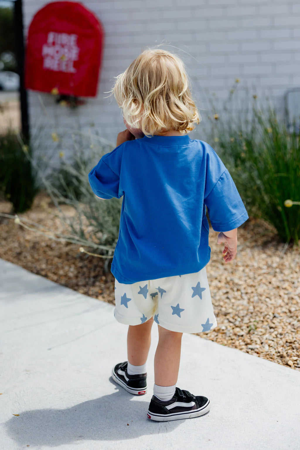 A young blonde child stands on the sidewalk, facing away, wearing an oversized Il Sole Tee by Sunday Siblings in blue/yellow and white shorts with blue stars. Black sneakers with white soles complete the outfit. A red mailbox and green shrubs are visible in the background.