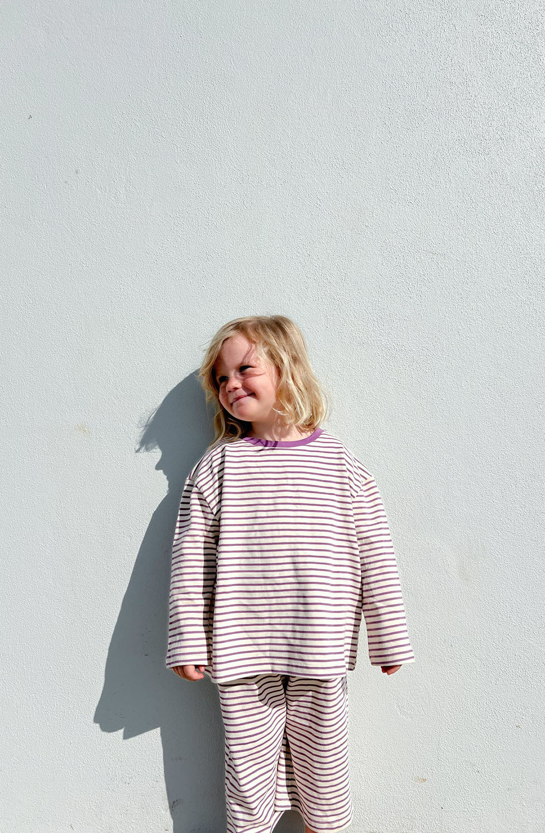 A young child with blonde hair stands against a plain white wall, wearing the Every Day Set Lavender from LENN LABEL, which consists of a matching striped long-sleeve shirt and pants made from 100% knit cotton. They are smiling with their eyes closed, leaning slightly to one side, enjoying the oversized fit and elastic waistband as they soak up the sunlight.