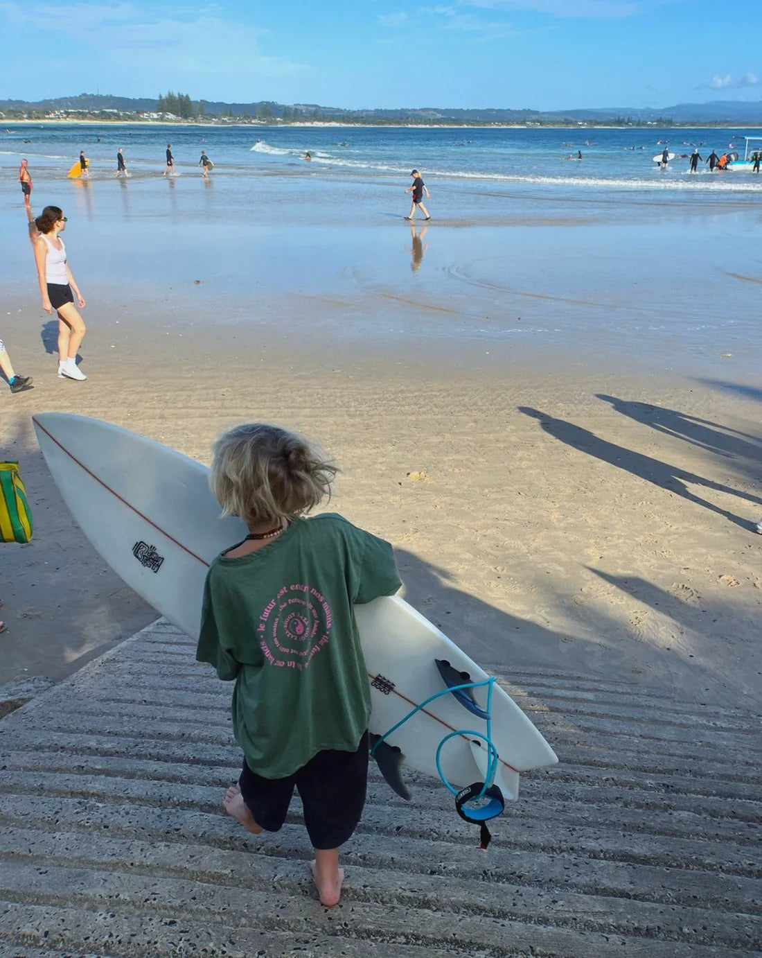 A child in a relaxed boxy fit Ziggy Zaza ~ Love Your Mother Tee Lizard walks down the steps towards a sandy beach, holding a white surfboard with a blue leash. Several people enjoy the water under a clear blue sky as shadows stretch across the steps.