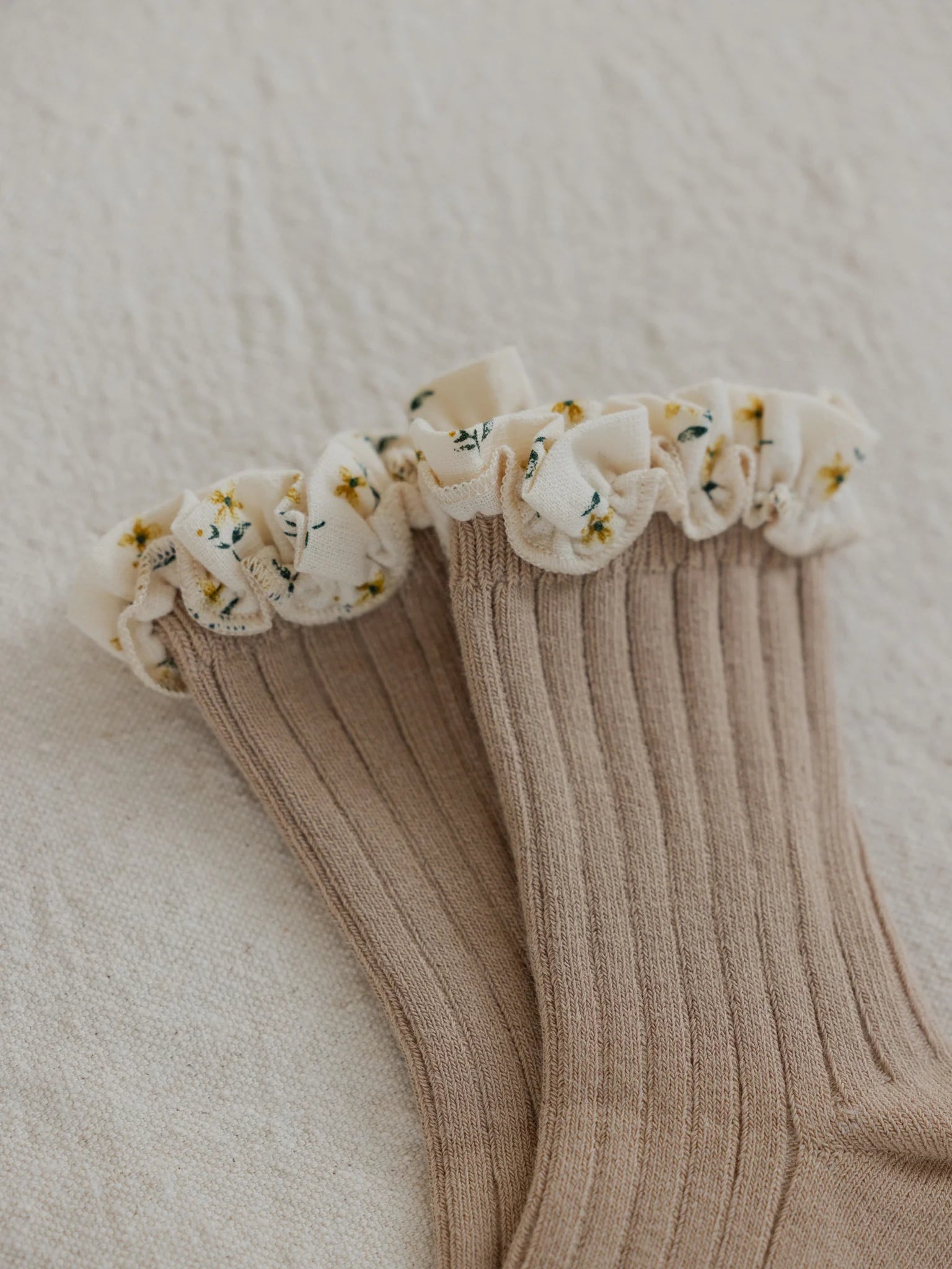 The Marigold Socks Clay by VALENCIA BYRON BAY are a pair of beige ribbed frilly socks featuring ruffled cuffs adorned with small yellow flowers and green leaves on a marigold printed frill, set against a light textured fabric background. Perfect for adding a touch of Byron Bay charm to your wardrobe.