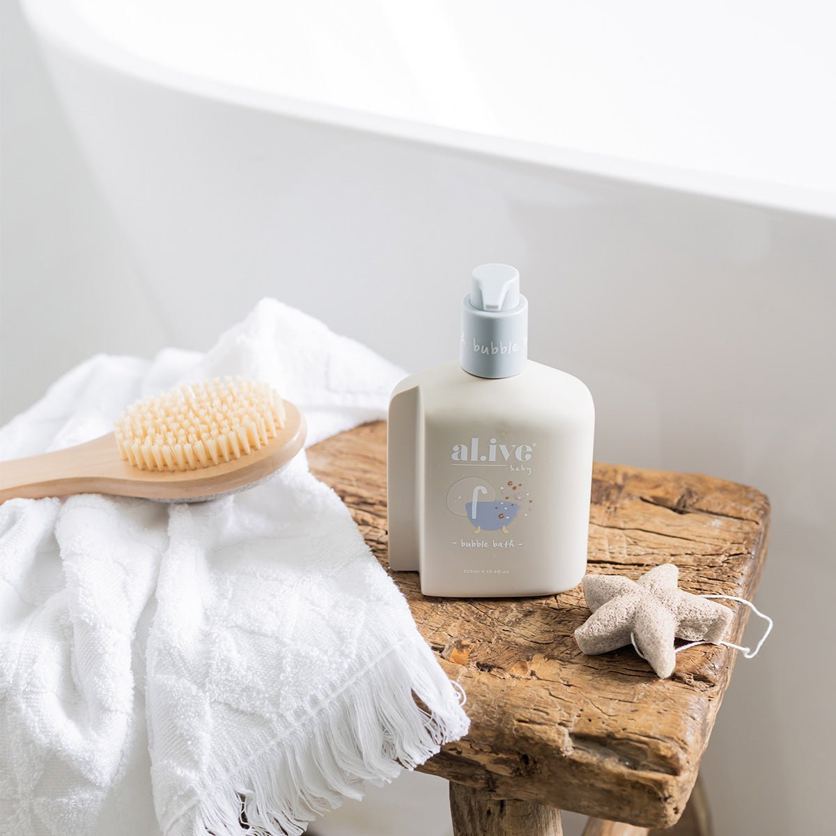 A bottle of AL.IVE bubble bath rests on a wooden stool beside a white towel and an AL.IVE BABY Konjac Sponge - White. A small starfish decoration is placed nearby, all set against the backdrop of a white bathtub.