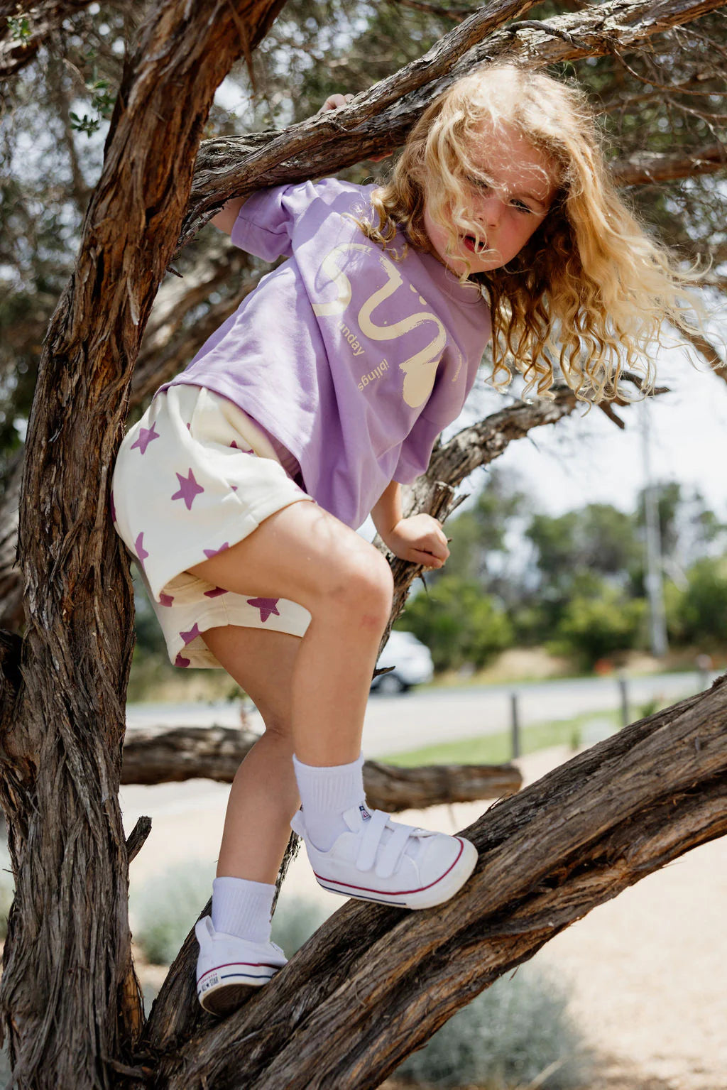 A young child with long curly hair climbs a tree outdoors, wearing a purple T-shirt and Sunday Siblings ~ Kiddo Shorts by SUNDAY SIBLINGS. The soft French terry cotton shorts have a lively star print, complemented by white socks and sneakers, with trees and a road in the background.