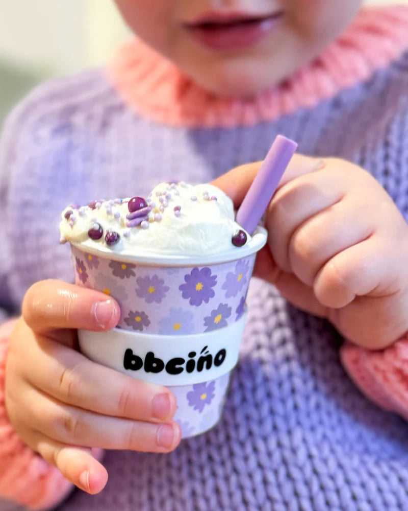 A child in a pastel-colored sweater holds the BBCINO Reusable Babycino Cup 120ml Blossom, an environmentally conscious cup designed with purple flowers and "bbcino" branding. This sustainable cup, crafted from bamboo fibers, is filled with whipped cream, topped with purple sprinkles, and comes with a matching straw.