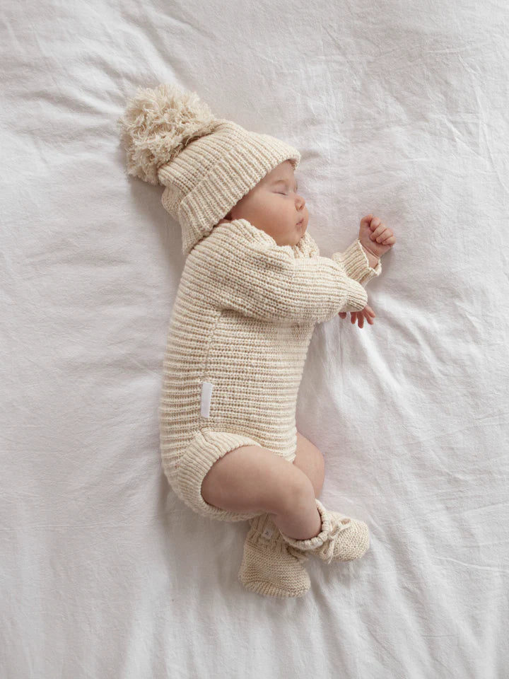 A sleeping baby is lying on a white bed, dressed in the cozy Heirloom Romper Honey from the ZIGGY LOU collection. The cream-colored outfit includes a hat with a pom-pom, a long-sleeved onesie, and booties. The baby has one arm slightly raised, resting peacefully on their side.