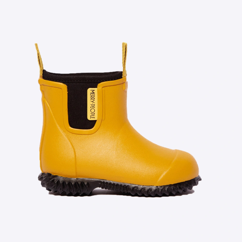 The Merry People Bobbi Kids Gumboot in mustard yellow features a black sole and lining, a back pull tab, side branding, and a wavy textured sole that is 100% waterproof. The product image has a plain white background.