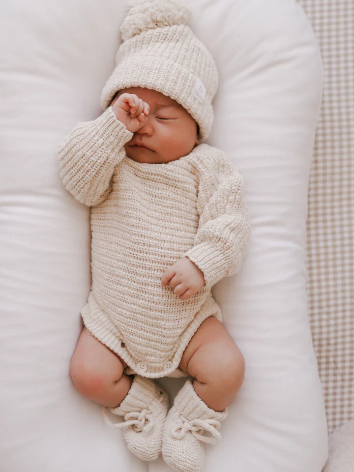 A baby is sleeping on a white cushioned surface, swaddled in the adorable Ziggy Lou Honey Signatures Collection. Sporting a knitted cream-colored Heirloom Romper Honey, a beanie with a pom-pom, and matching booties from ZIGGY LOU, the baby has one hand lightly covering their face.