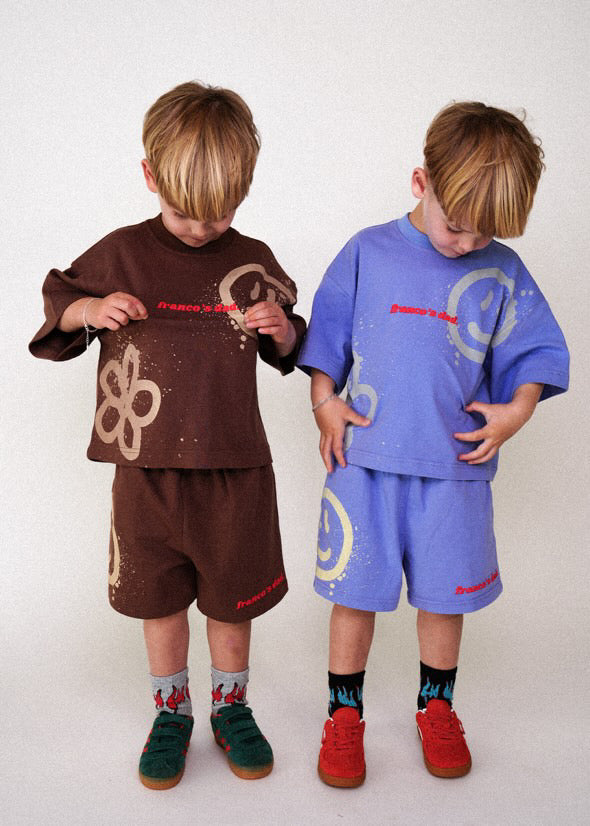 Two young children stand side by side in stretch cotton lounge sets. One wears Franco's Dad Acid Dye Shorts Brown with flower designs; the other has a blue outfit with smiley faces, both having blonde hair and red text accents, looking down at their clothes.