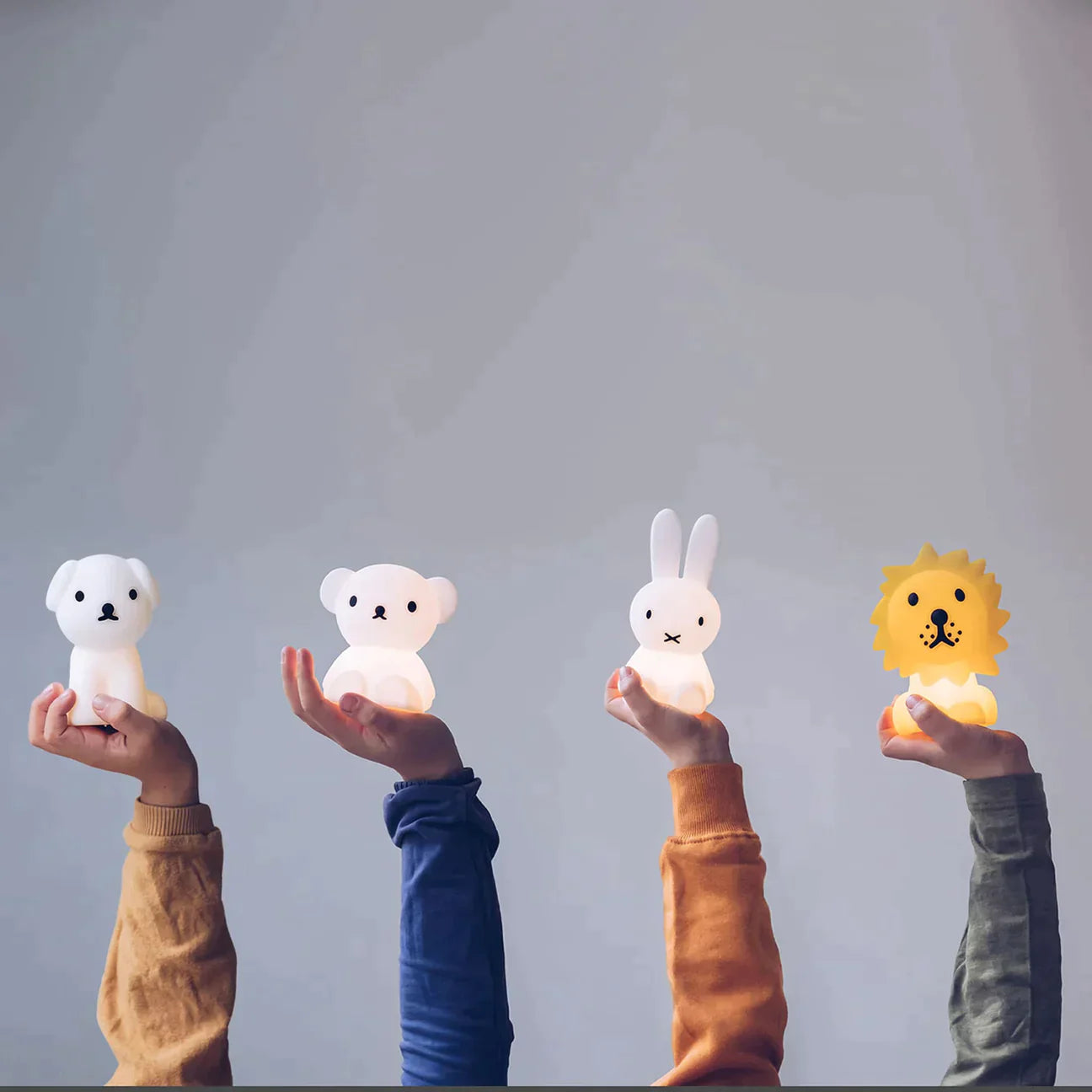 Four hands hold up different animal-shaped nightlights from MR MARIA's Bundle of Light - Miffy collection against a plain background. From left to right: a white dog, a white bear, a white bunny, and a yellow lion—all soft silicone LED lamps. Each hand is adorned with differently colored sleeves (orange, blue, mustard, and burgundy). These make perfect gift display items.