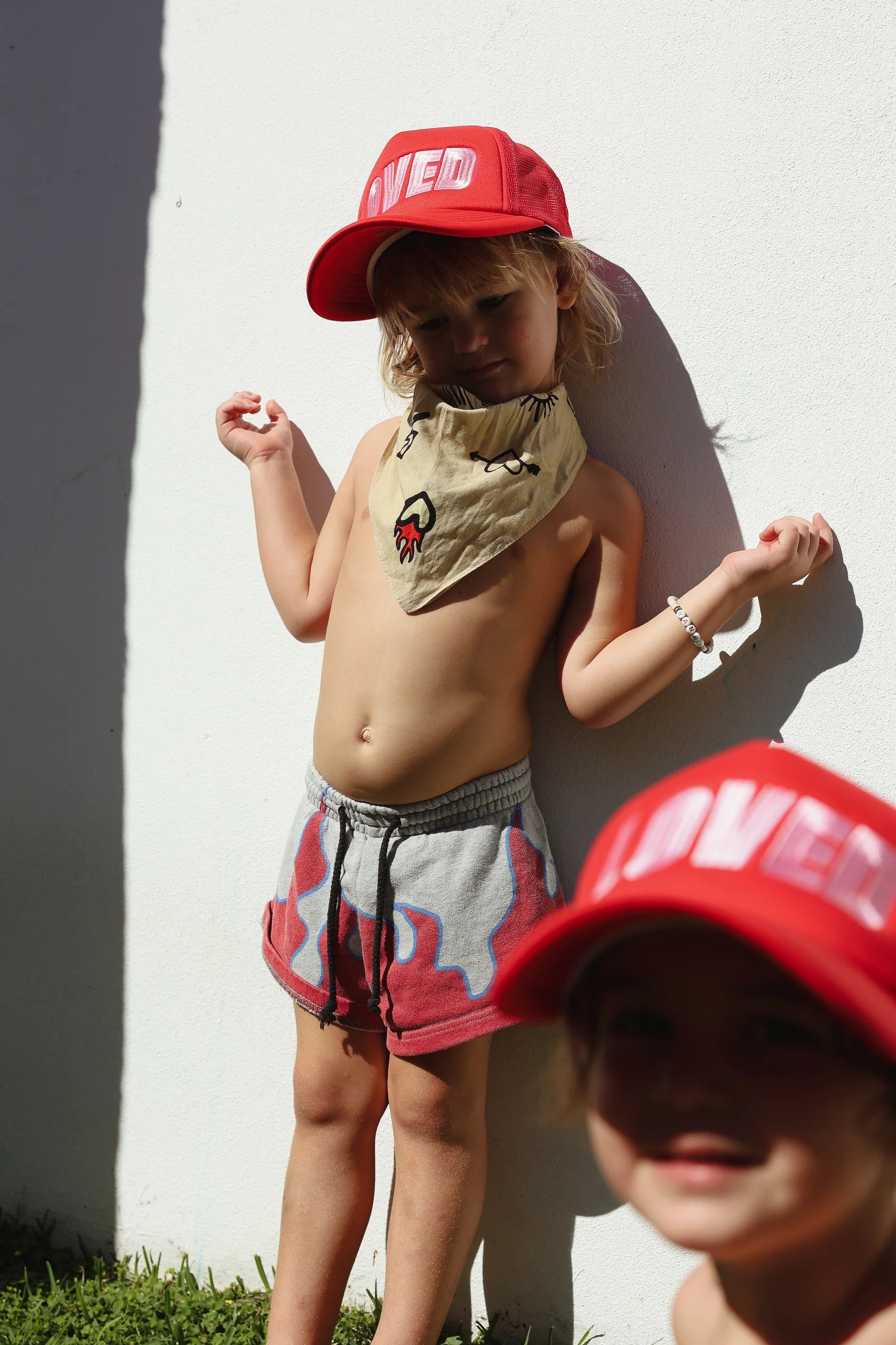 Two young children are outdoors against a white wall. The child in the foreground wears a red "So Loved Trucker" cap from TINY LOVE CLUB with an adjustable back strap, paired with a beige neck scarf and shorts. Another child, also wearing the sun-safe red "So Loved Trucker" cap from TINY LOVE CLUB, smiles at the camera.