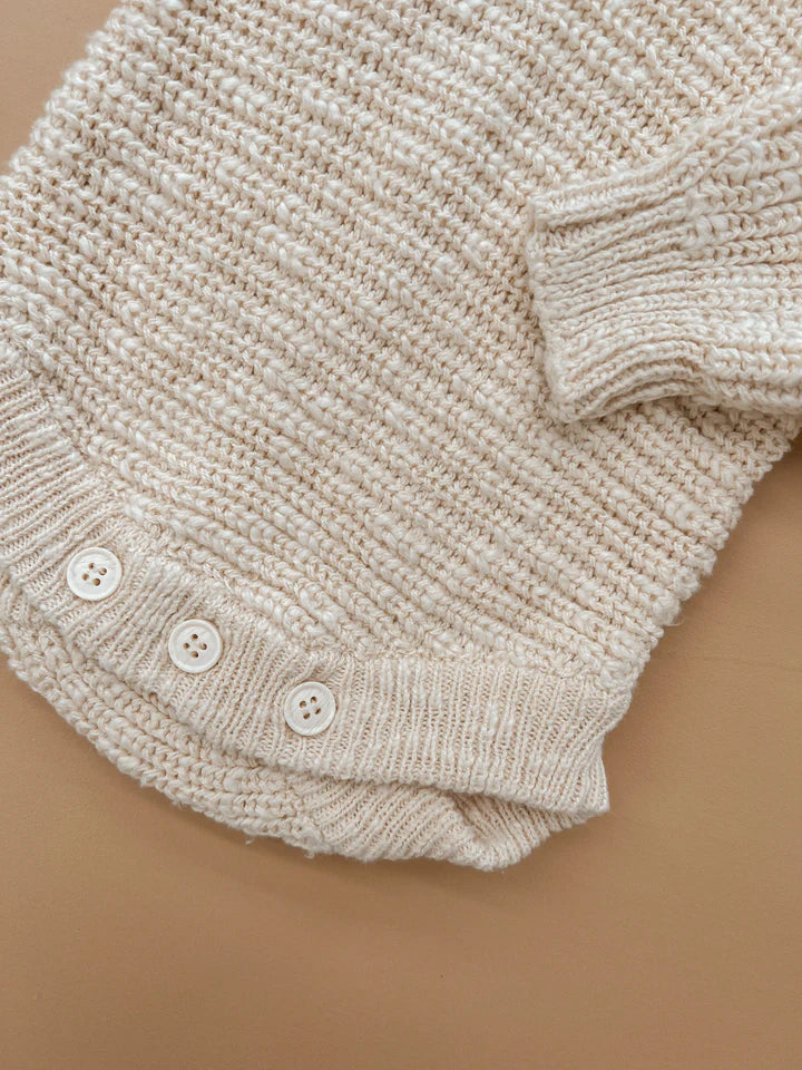 A close-up of a sleeve from the Heirloom Romper Honey by ZIGGY LOU, showcasing three white buttons near the cuff against a light brown backdrop. The beige, textured knit pattern is clearly visible, emphasizing the romper's cozy and detailed craftsmanship.