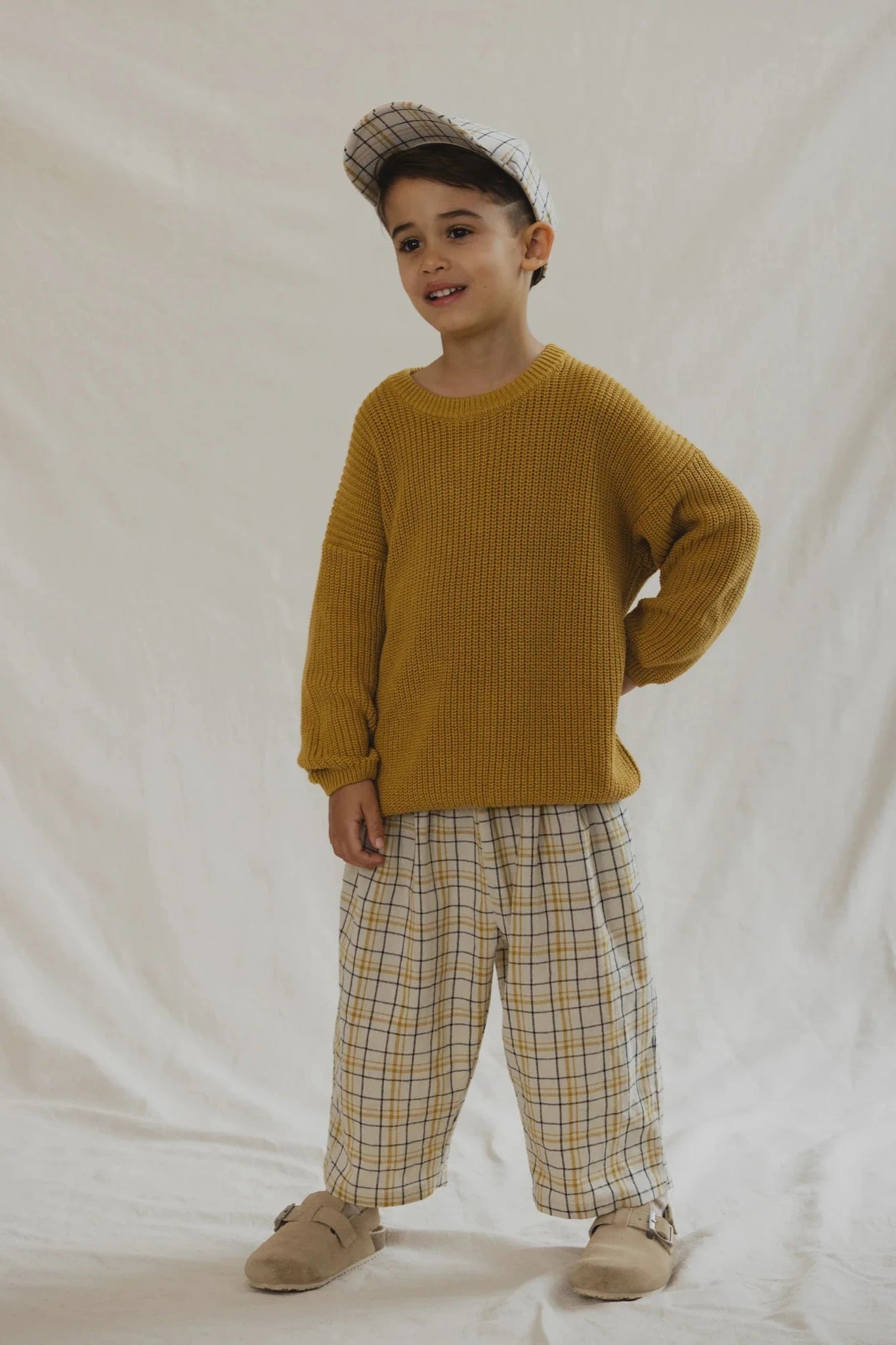 A child in a mustard yellow sweater, Rio Pant Picnic plaid pants with an elastic waistband, and a matching plaid cap by VALENCIA BYRON BAY stands against a plain backdrop, smiling with one hand on their hip and highlighting this unisex outfit's versatility.