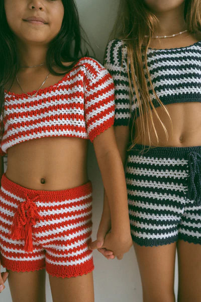 Two young girls hold hands, wearing NOTHING BUT AMOR's handmade Crochet Stripes Sets. One wears red/white and the other black/white outfits, both crafted from soft cotton with short-sleeve tops and matching shorts, exuding Bali's charm.