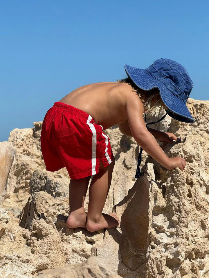 A young child in red shorts, wearing the ZIGGY ZAZA 'Ziggy Zaza ~ Everyday Scout Hat Blu' with an adjustable chin strap, is climbing beige rocks under a clear sky. The cotton canvas hat fits snugly as the barefoot child determinedly explores the rocky terrain, facing away.