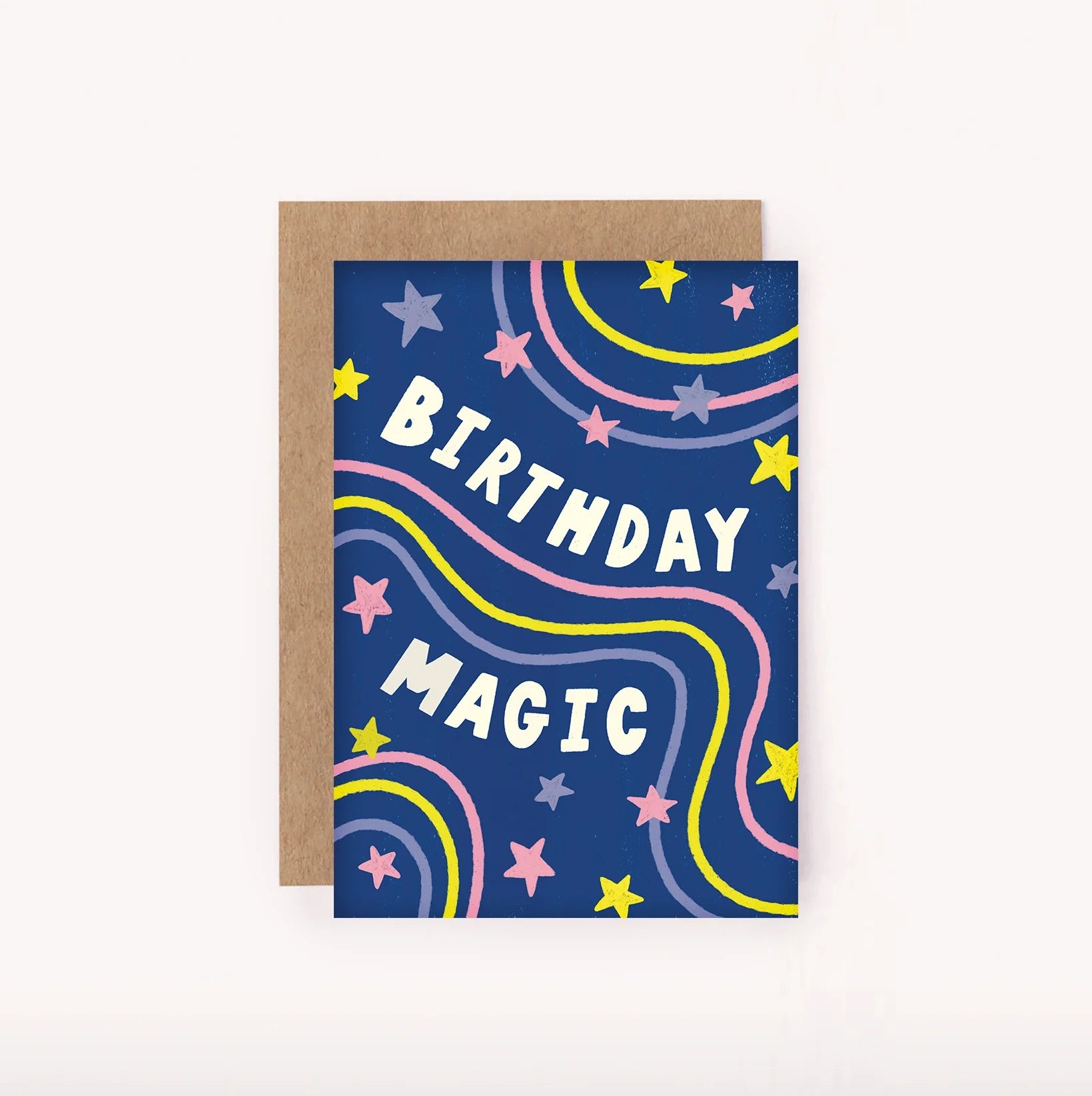 The "Birthday Magic Mini Card" by LAUREN SISSONS STUDIO features white text reading "Birthday Magic" on a dark blue background, made from recycled paper. The card is adorned with colorful stars and wavy lines in pink, yellow, and green. Behind the card lies a brown envelope.