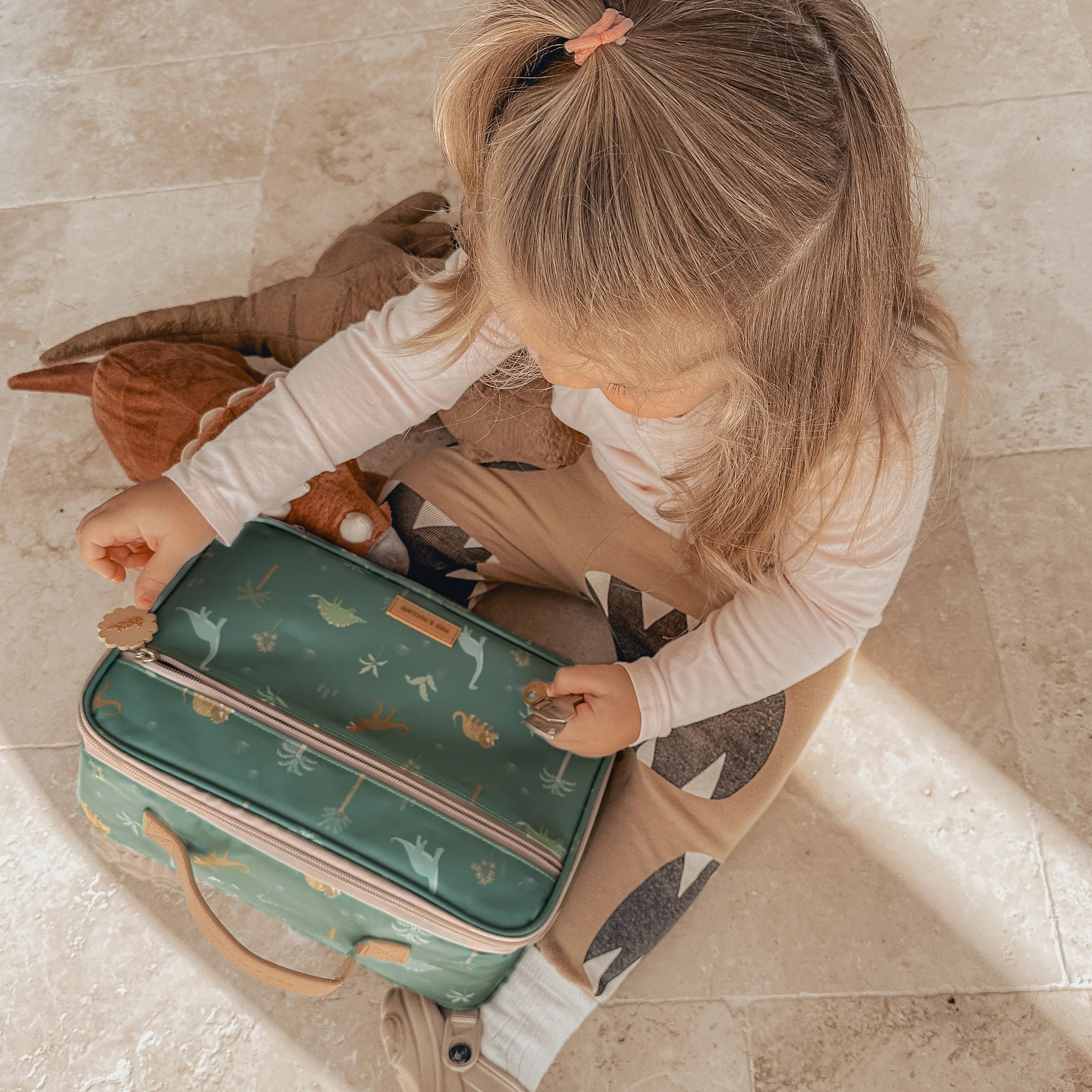 A young child with long blonde hair is sitting on the tiled floor holding a small, green suitcase adorned with animal illustrations. The child, dressed in beige pants and a white top, has a FOX & FALLOW Dinos Lunch Bag beside them along with a plush toy for company.