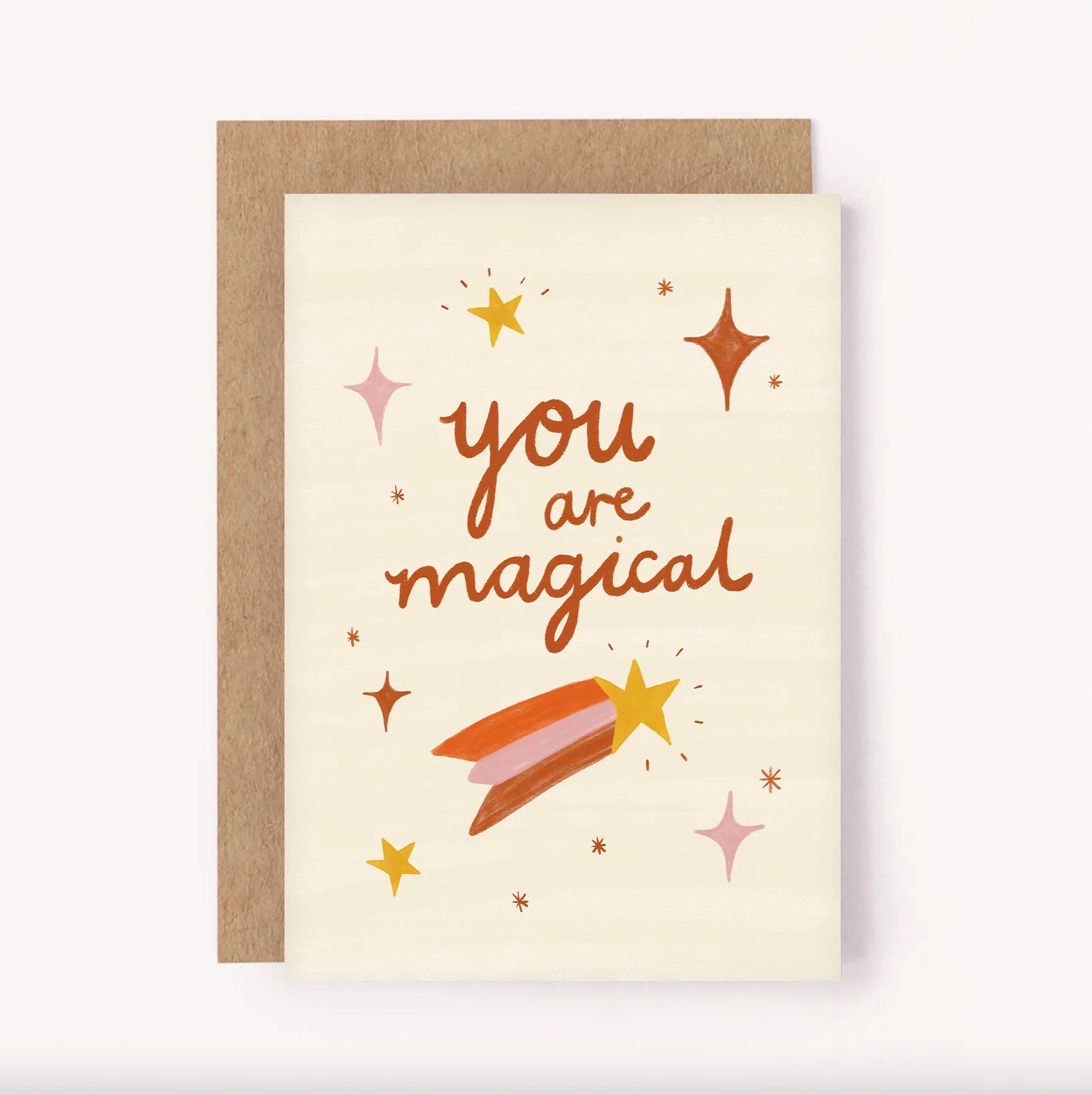 The LAUREN SISSONS STUDIO Greeting Card "You Are Magical" is crafted from recycled paper and features a beige background with a brown envelope. Adorning the card in brown lettering is the message "You are Magical," embellished with gold, pink, and brown stars and sparkles. The enchanting design is completed by a shooting star with a gold tail.