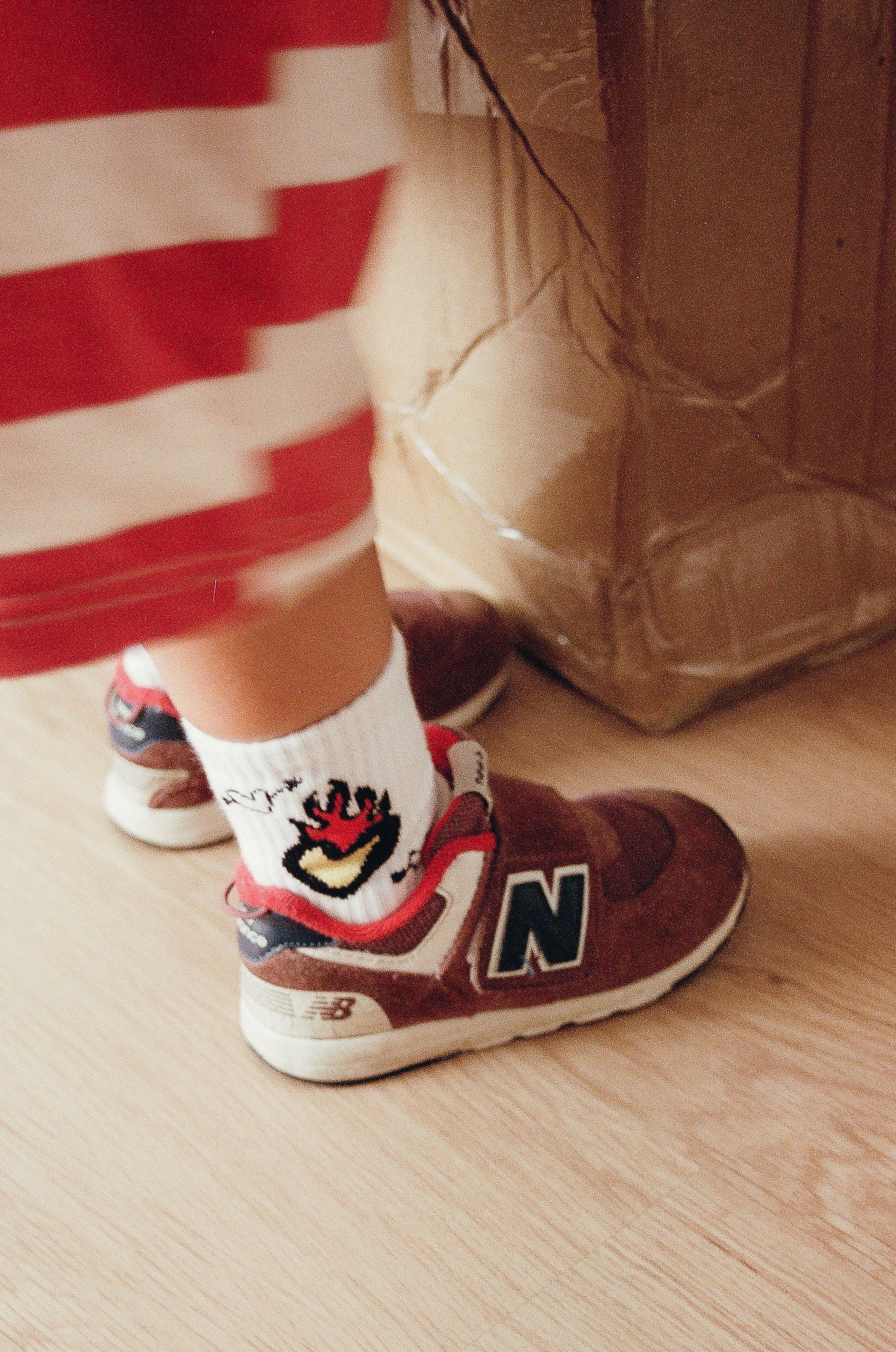 Close-up of a child's feet, aged 2-5 years, sporting red and white sneakers with an "N" logo, tiny Burning Love Socks by TINY LOVE CLUB featuring a playful rooster design, and red and white striped pants. The child stands next to a large, taped cardboard box on a wooden floor, perfectly capturing their sports style.