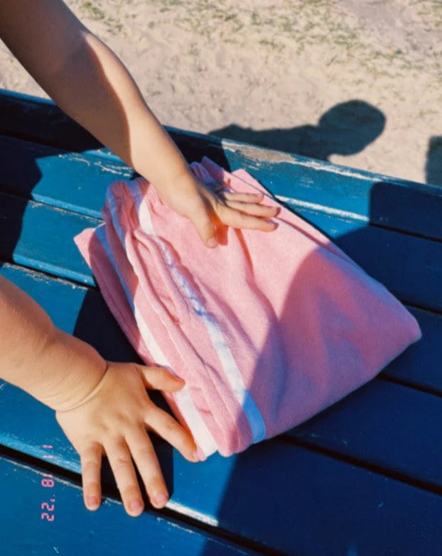 A child's hands touch the Ziggy Zaza ~ Twin Stripe Terry Pant Pink by ZIGGY ZAZA on a blue wooden surface, with shadows dancing and sand and grass in the background.