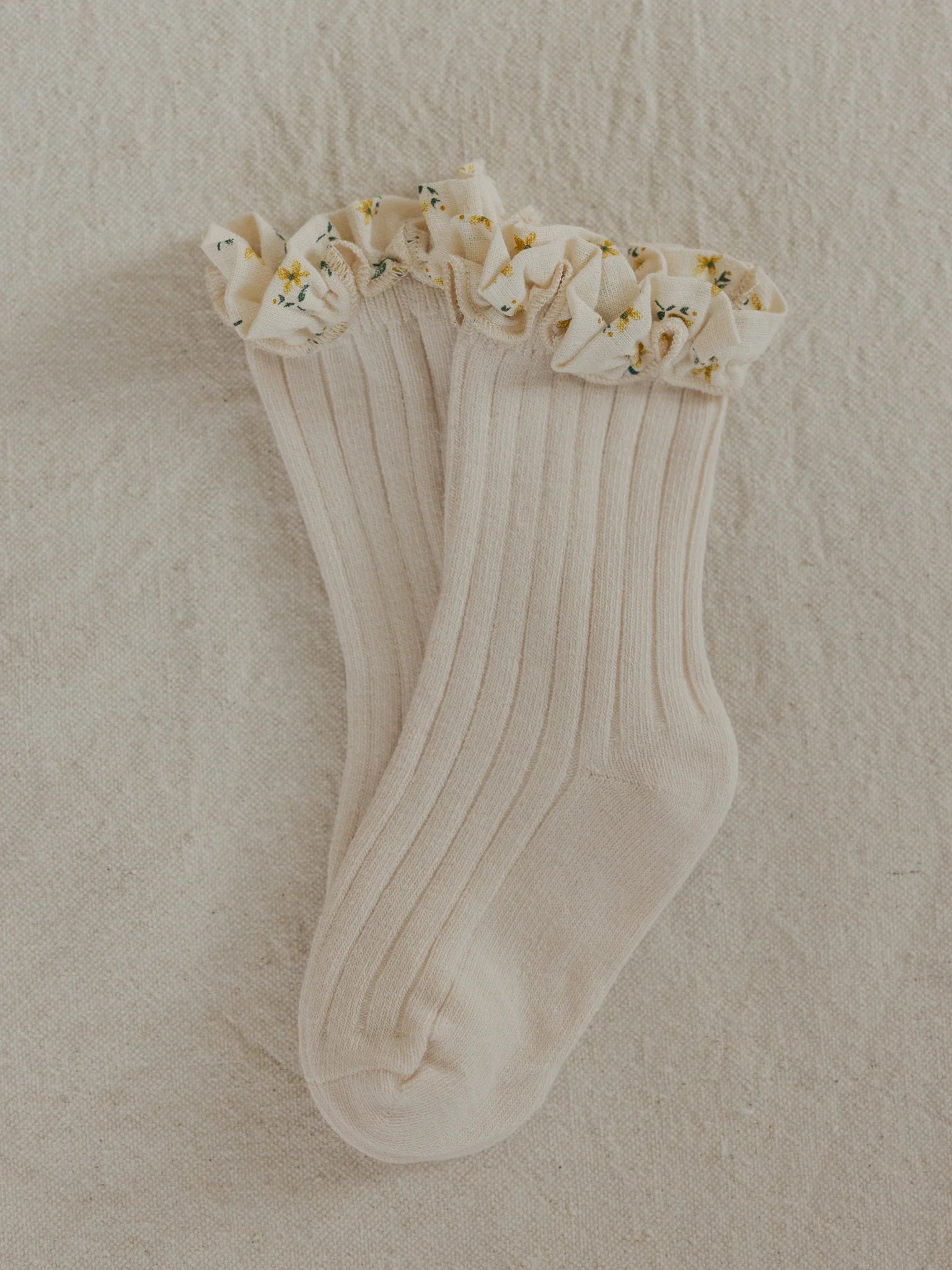 The Marigold Socks Sand by VALENCIA BYRON BAY are ribbed, cream-colored socks with a frilly trim. The marigold-printed frill around the top features small yellow flowers and green leaves, and they are displayed lying flat on a textured light beige surface.