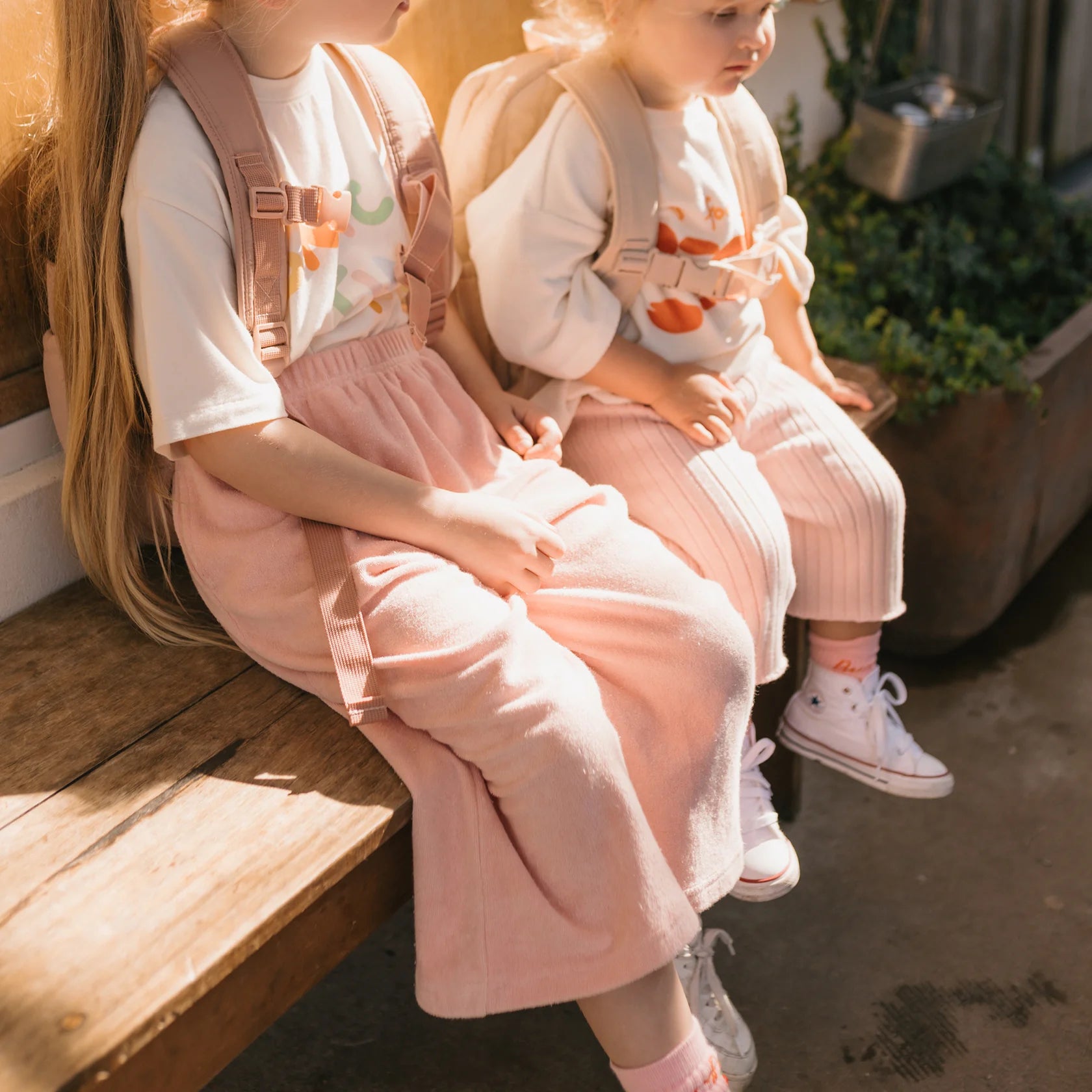 Two young girls sit on a wooden bench, both wearing white shirts and light pink overalls; one pair features long pants while the other sports GOLDEN CHILDREN Confetti Wide Leg Pants in a delightful pink parfait tone. Light-colored shoes and backpacks complete their outfits as sunlight casts gentle shadows on the ground.