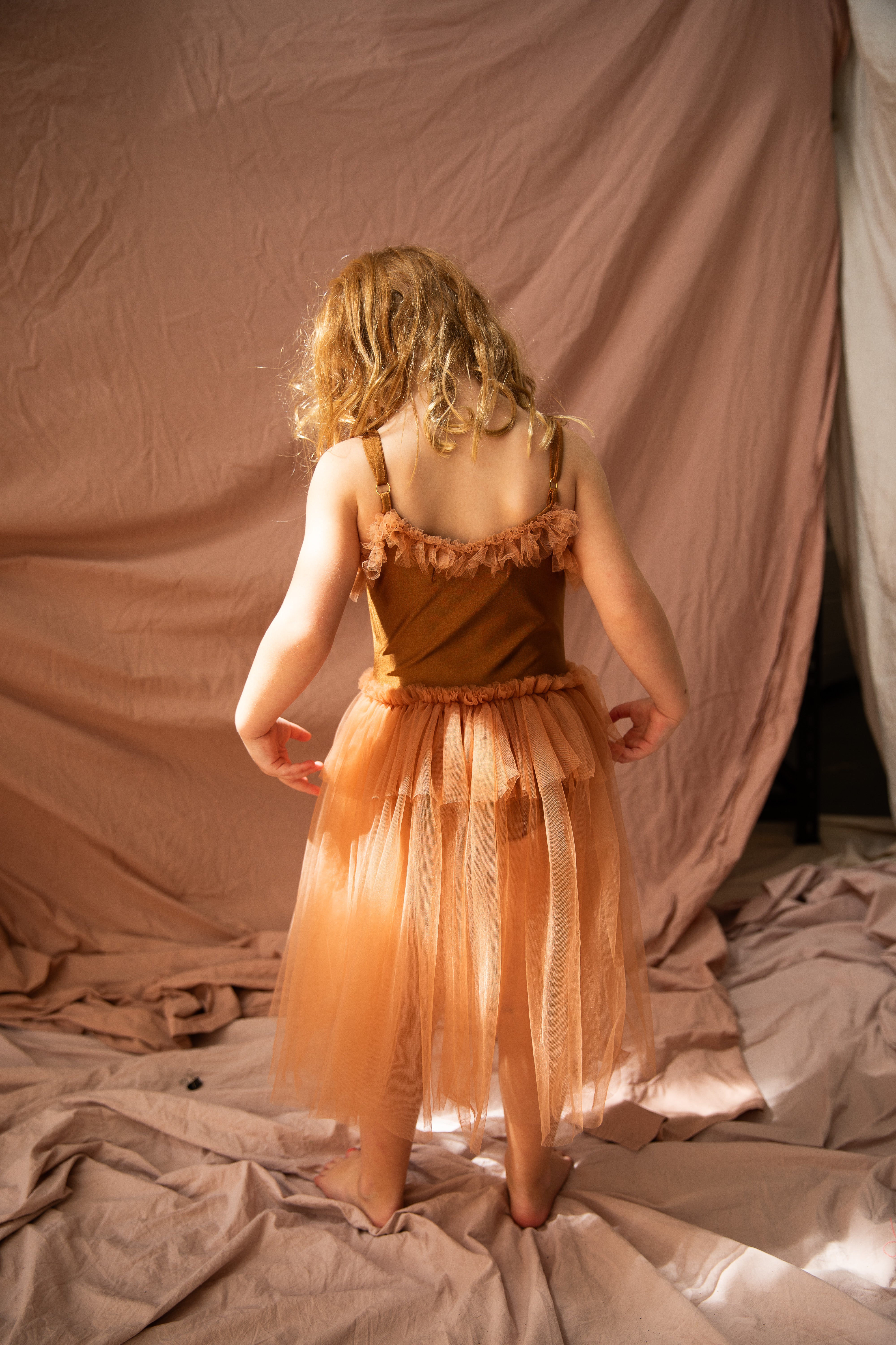 A young child with curly blonde hair stands barefoot on a fabric-covered floor, wearing the Bella + Lace Elanore Dress in Coffee Bean (Size 1-2). The ruffled peach tutu and draped beige-pink backdrop evoke a ballet class. The child faces away from the camera.