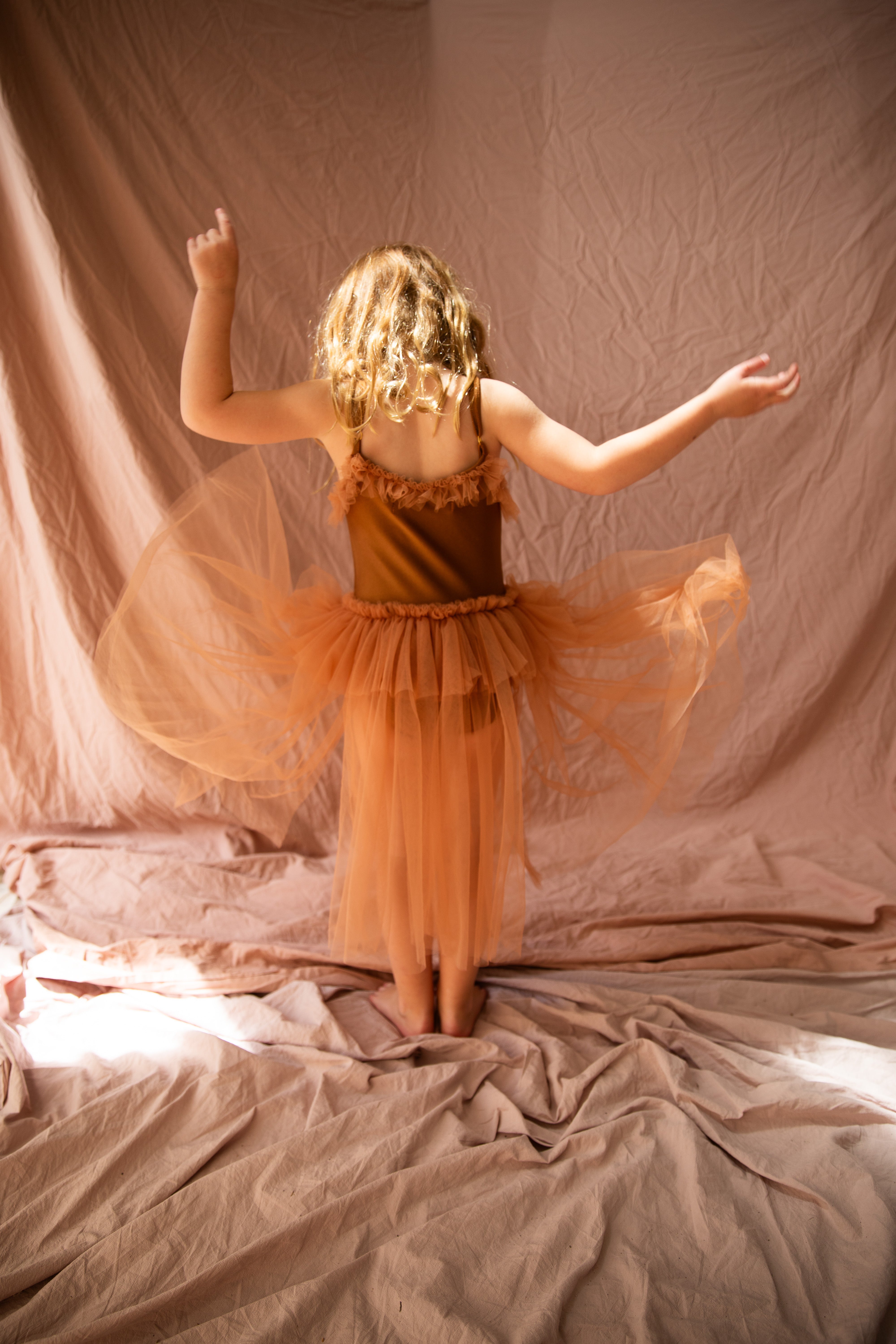 A young child dressed in the Elanore Dress Coffee Bean by BELLA + LACE twirls with arms raised in a softly lit room. The backdrop of delicate tulle and flowing beige fabric creates a dreamy atmosphere, evoking the charm of an enchanting ballet class.