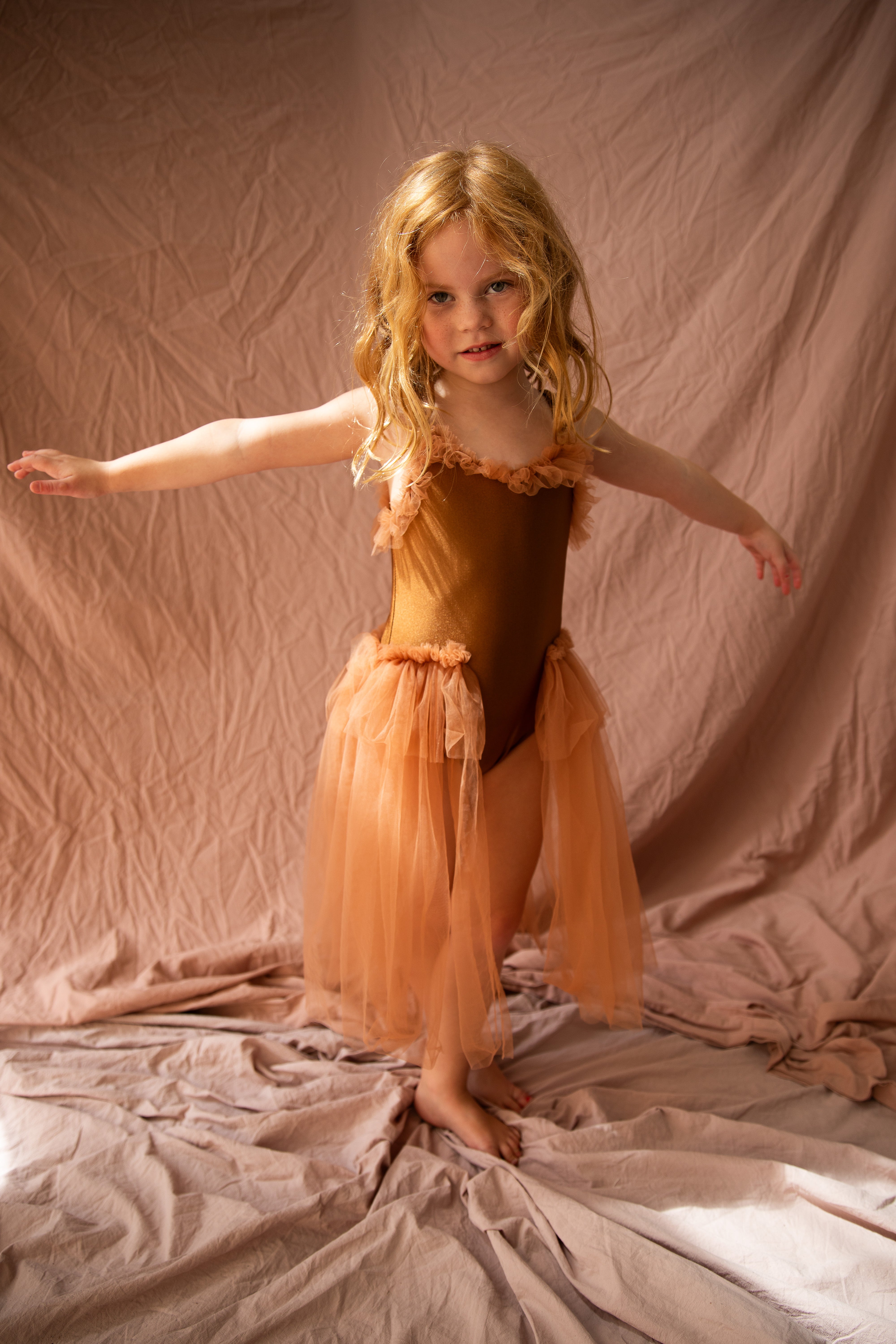 A young girl with long blonde hair stands barefoot on a soft, light-colored fabric, wearing the BELLA + LACE Elanore Dress Coffee Bean (Size 1-2) with soft tulle accents. She holds her arms outstretched against a matching backdrop.