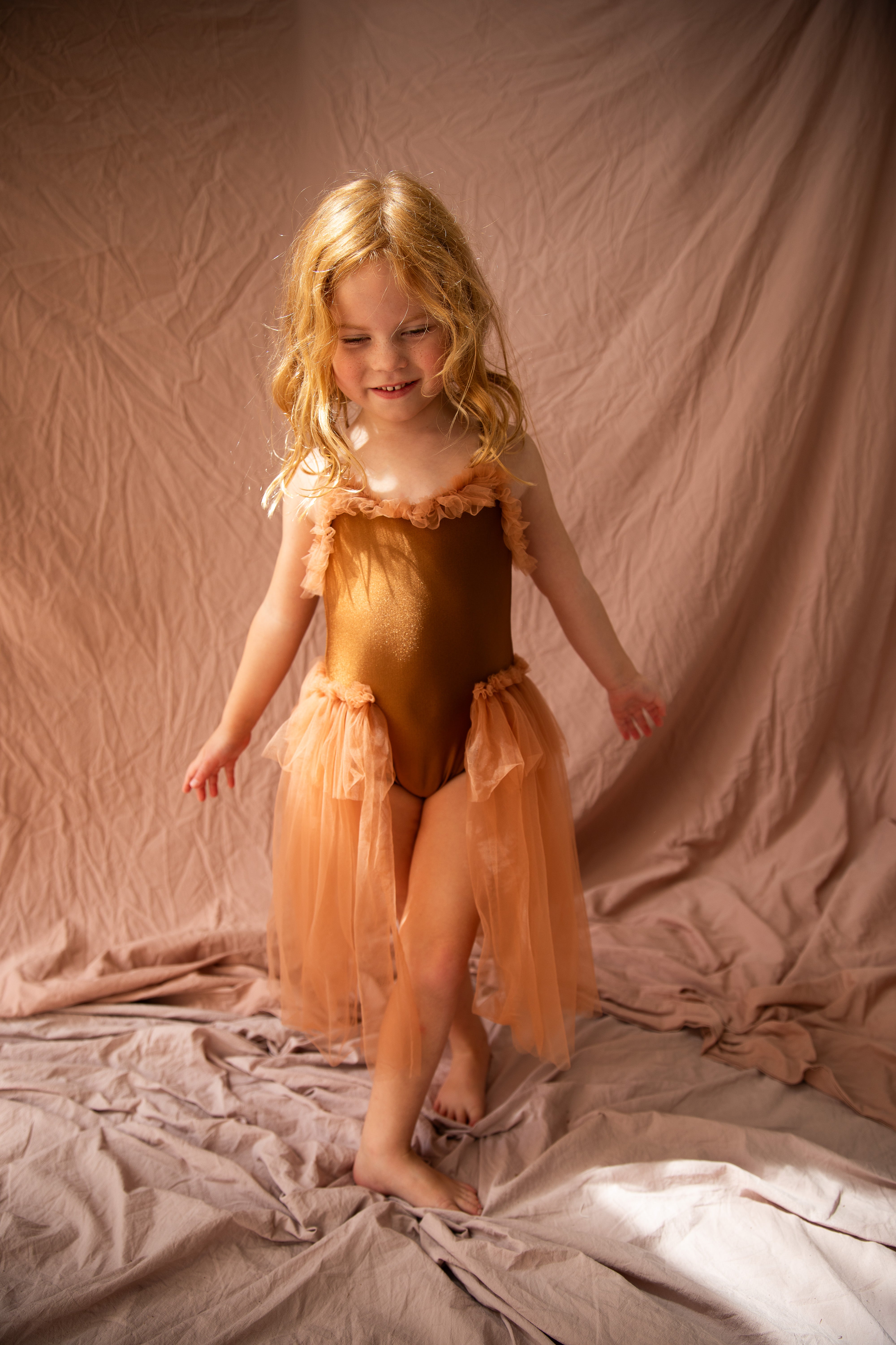 A young girl with blonde hair beams in her Elanore Dress Coffee Bean by BELLA + LACE against a soft pink draped background. She looks joyful and playful, ready for her ballet class.