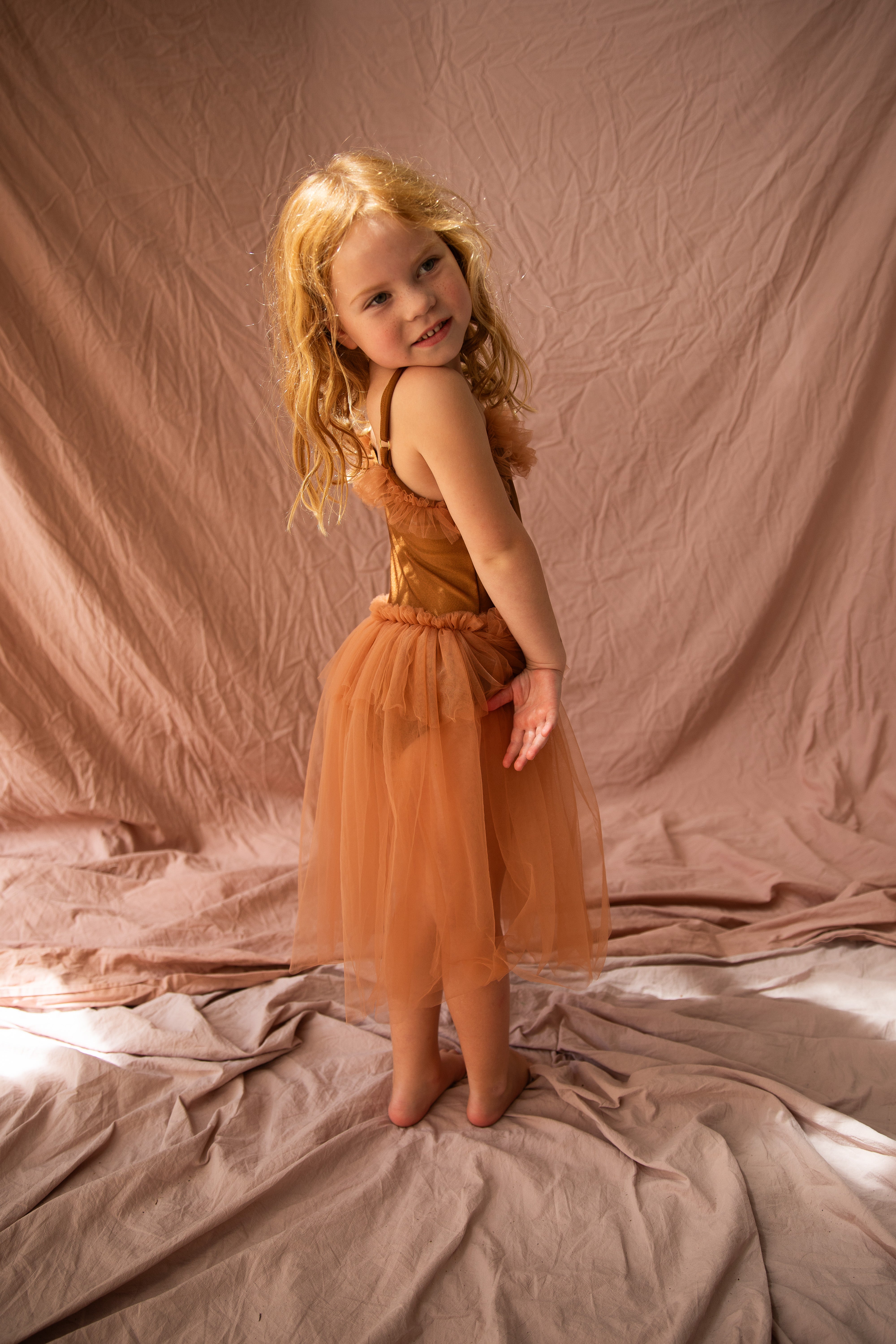 A young girl with curly blonde hair playfully poses in the BELLA + LACE Elanore Dress Coffee Bean, featuring a soft tulle skirt, against a backdrop of draped beige fabric.