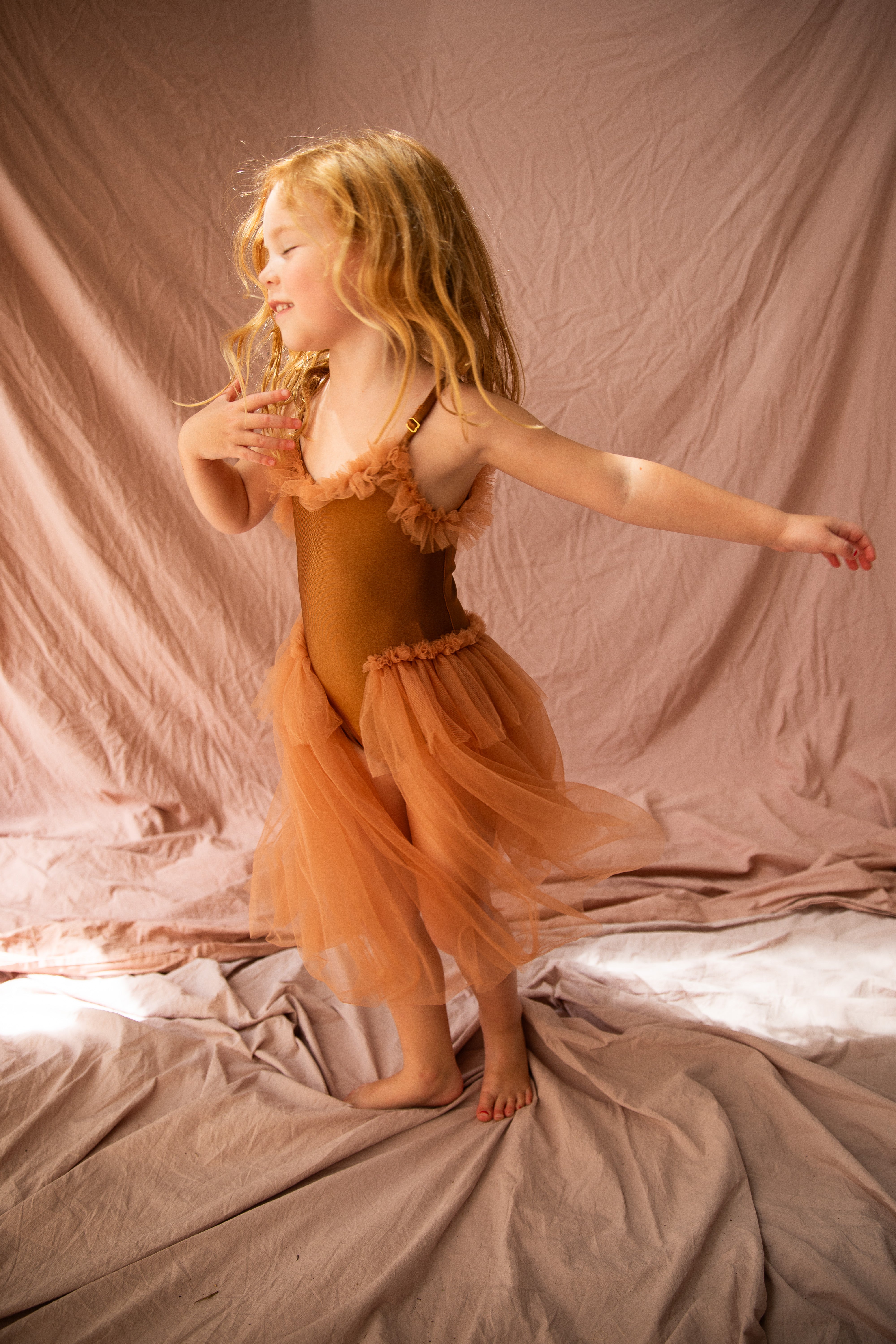 A young girl with long blonde hair joyfully dances on a beige fabric surface, wearing the BELLA + LACE Elanore Dress Coffee Bean in Size 1-2. Her eyes are closed as she moves gracefully, reminiscent of a ballet class, against a matching beige background.