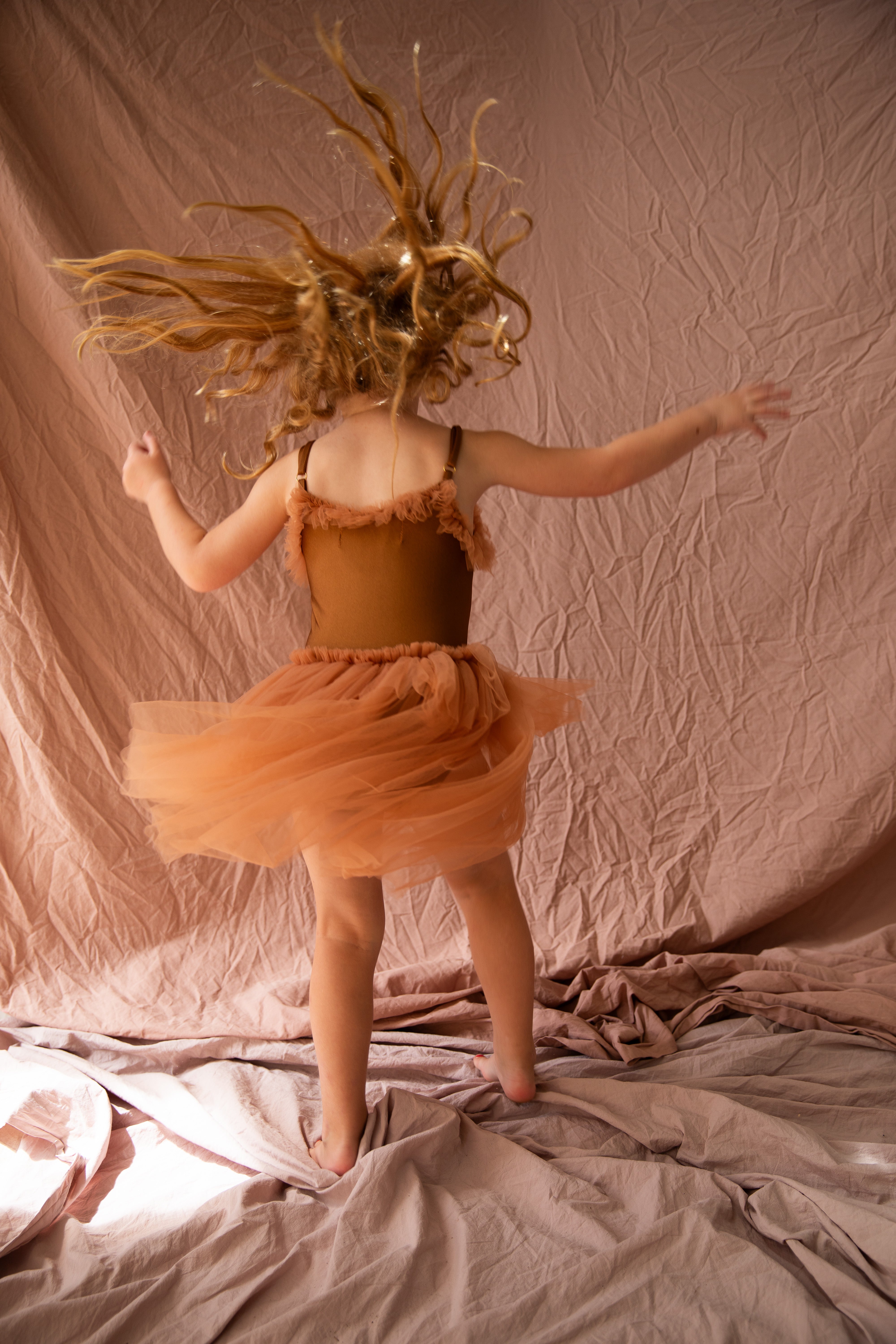 A young girl twirls in a BELLA + LACE Elanore Dress Coffee Bean - Size 1-2, on a crinkled fabric floor, hair flying with motion. The matching brown background enhances the ballet-like grace.