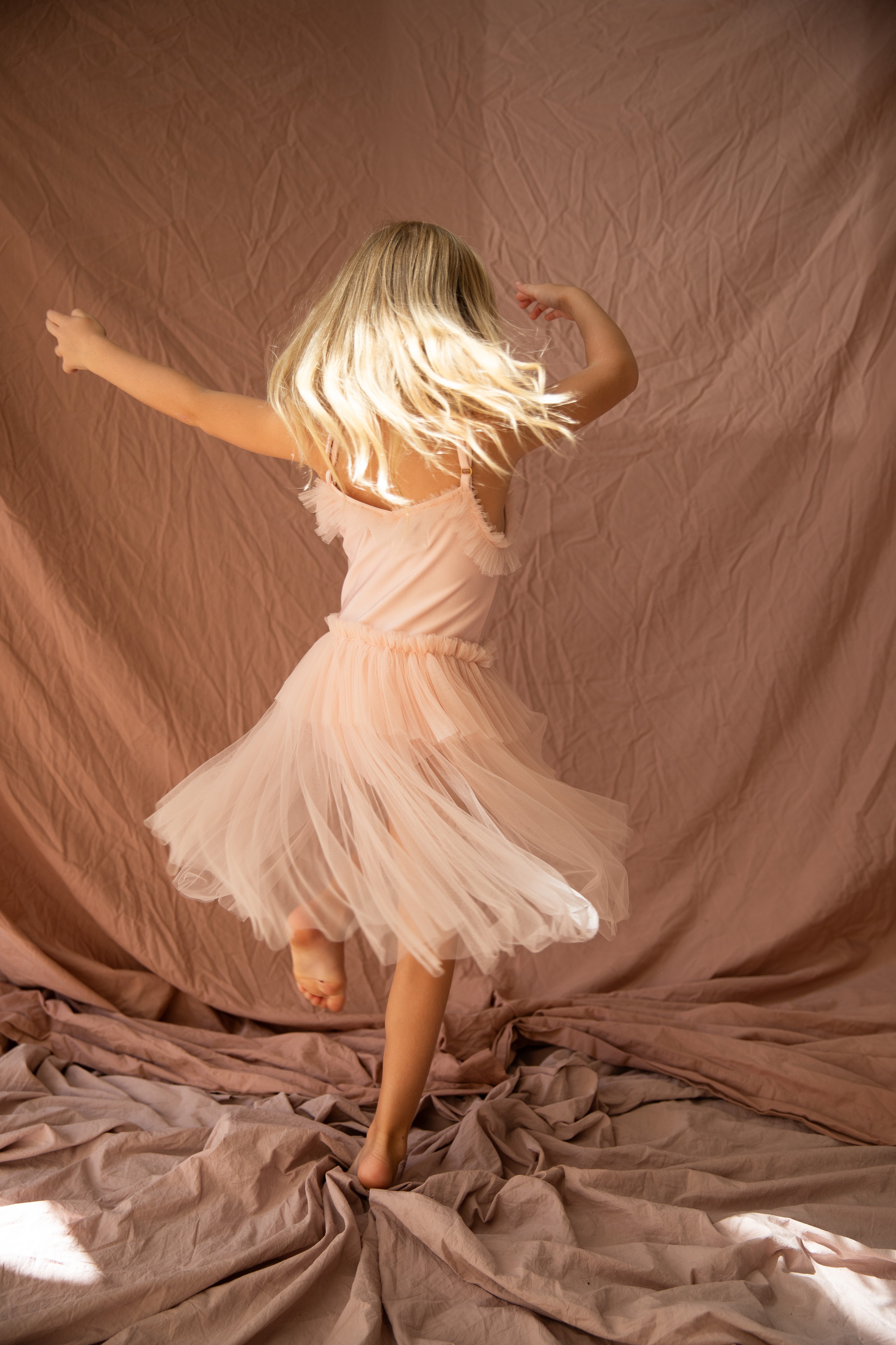 A young girl named Elanore with long blonde hair joyfully twirls in the BELLA + LACE Elanore Dress Twinkle Toes, crafted from pale pink tulle. Dancing on soft, crumpled fabric with a matching backdrop reminiscent of her ballet class, she creates a whimsical and dreamy scene.